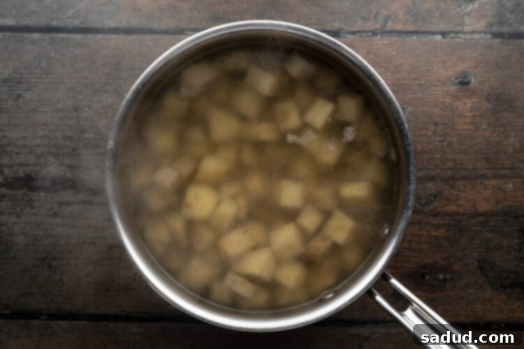 Overhead view of large silver pot holding cubed potatoes and cold salted water sitting on a wooden tabletop, ready to be boiled.