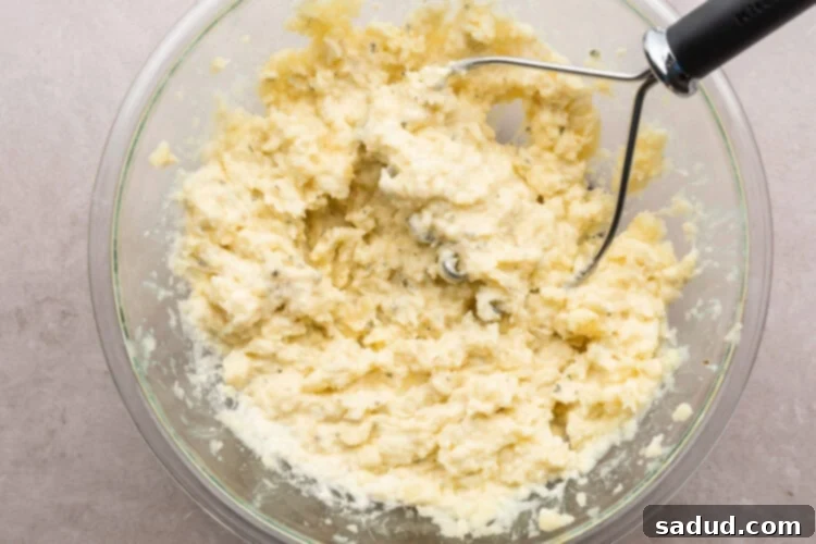 Overhead view of creamy mashed potatoes in a large glass mixing bowl with a potato masher resting on a neutral countertop.
