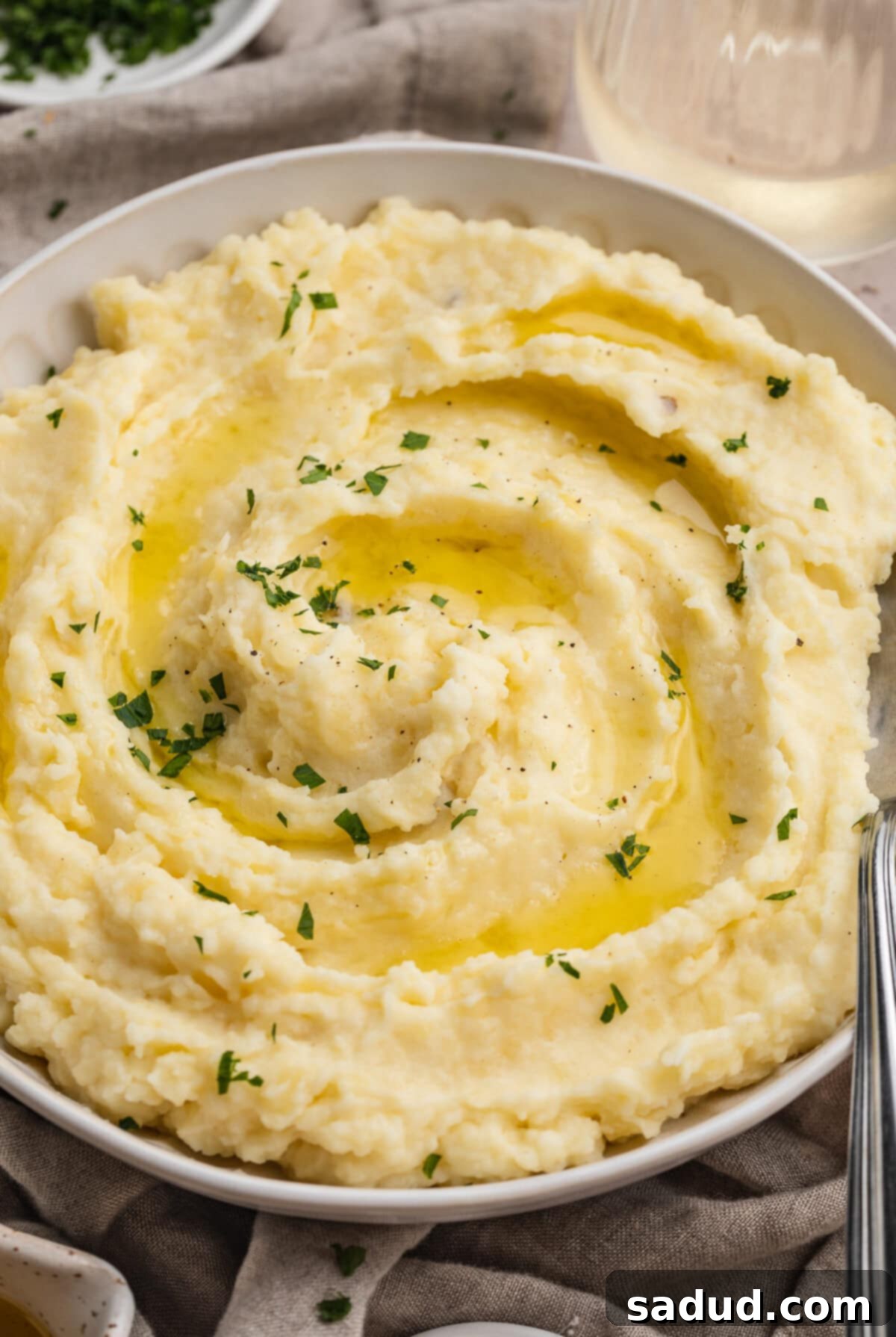 Angled look at a bowl of truffle mashed potatoes, topped with truffle oil and chopped parsley, in a large bowl on a dining table, with a serving spoon.