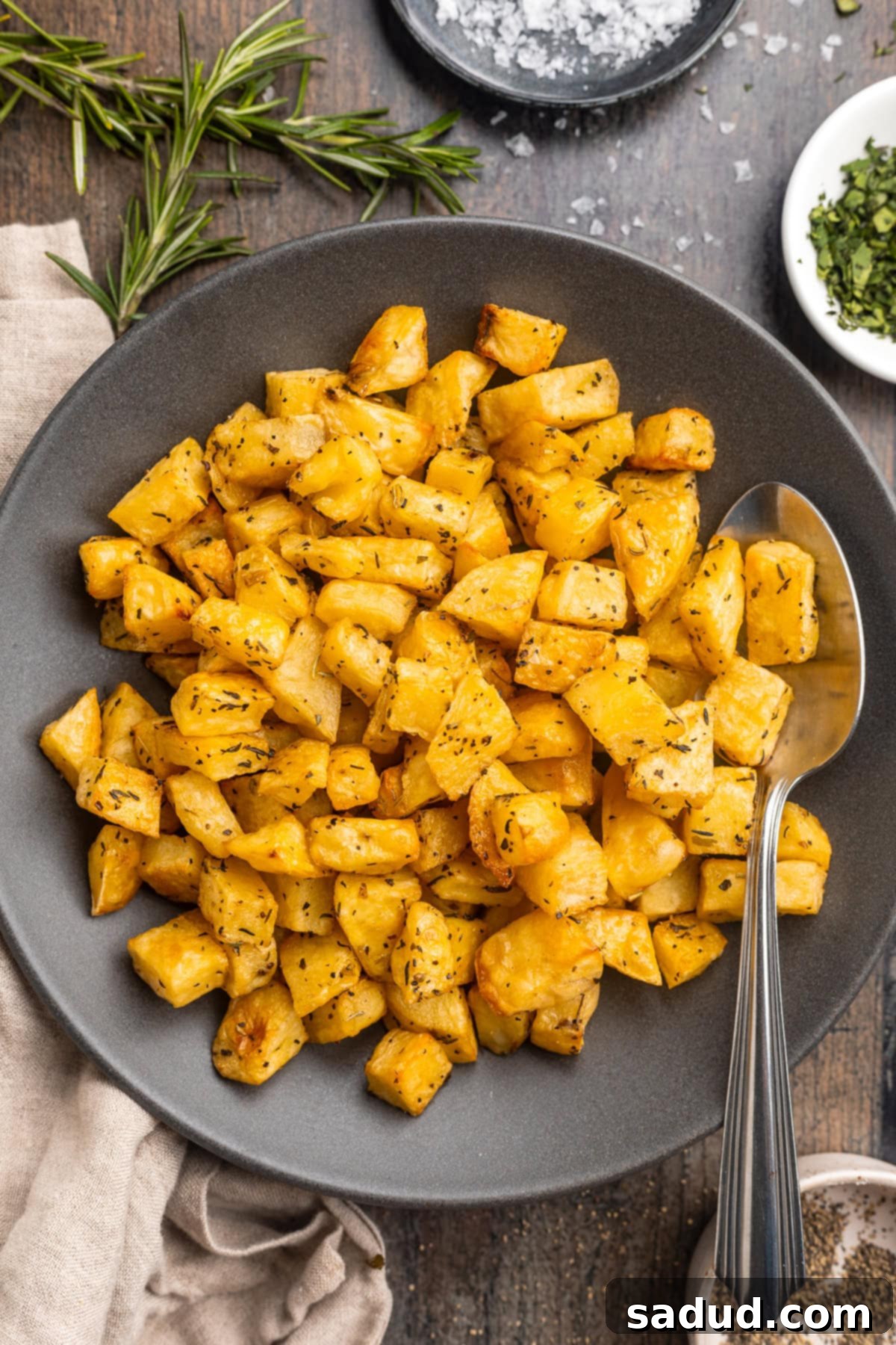 Close-up overhead view of raw diced potatoes seasoned generously with dried herbs, salt, and pepper in a rustic wooden bowl on a wooden table.
