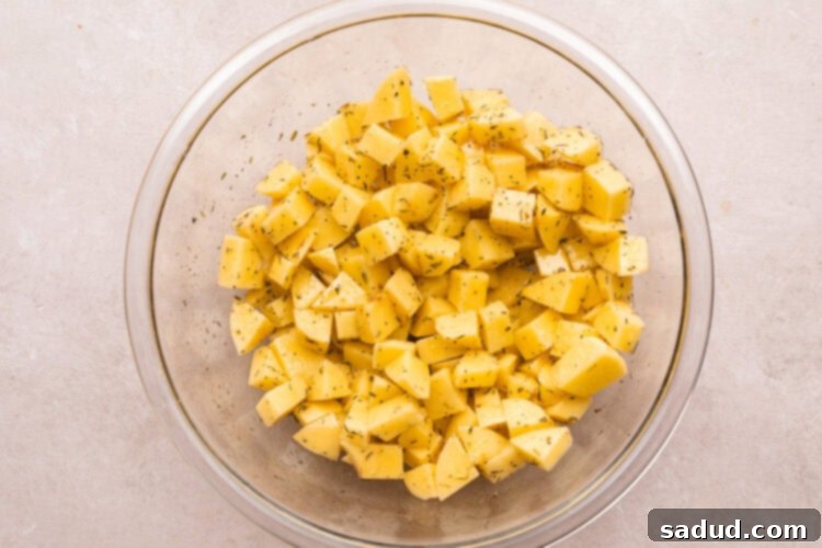 Overhead view of diced potatoes in a large glass mixing bowl, being tossed with oil and seasoning.