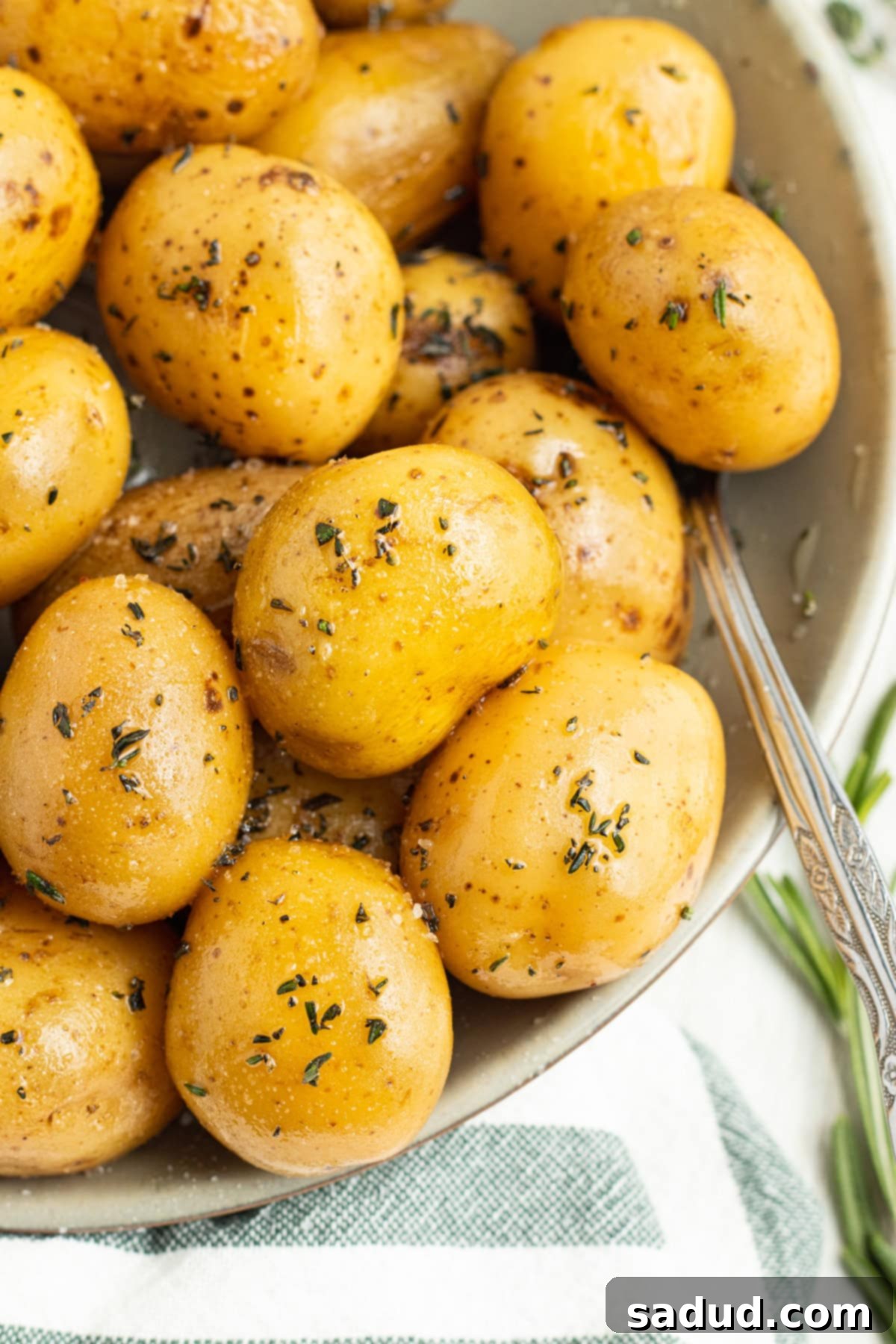 Overhead, close-up view of golden confit potatoes in a large white bowl on a white tablecloth, garnished with fresh herbs.