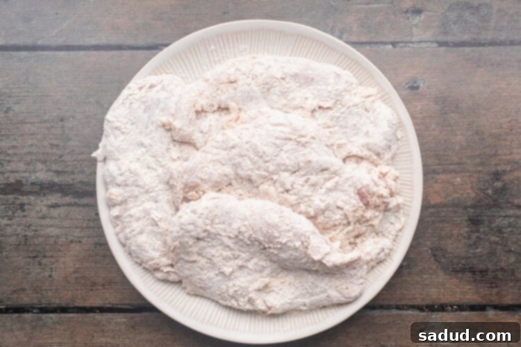Breaded chicken breasts on a round white plate, ready for frying.