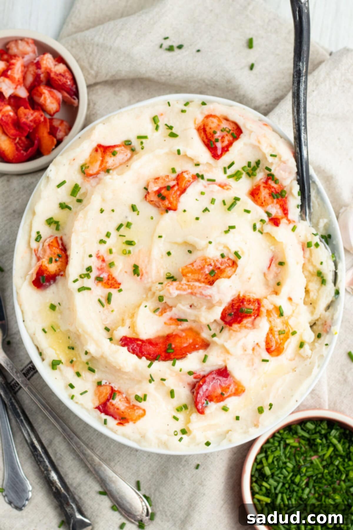 Top-down view of a giant bowl of swirled, whipped mashed potatoes with large chunks of pink and red lobster meat, garnished with fresh herbs.
