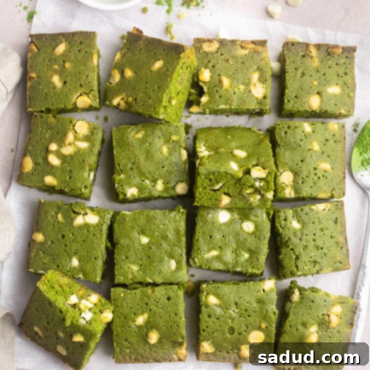 Matcha Magic White Chocolate Brownies 4 Squares of bright green matcha brownies peppered with white chocolate chips stacked up on parchment paper on a countertop.