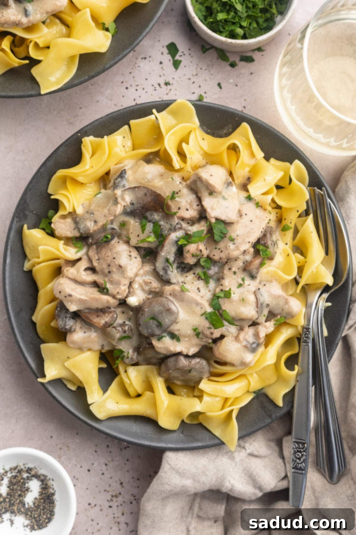 Zoomed out overhead photo of savory pork stroganoff in a dark grey bowl with egg noodles and fresh herbs.