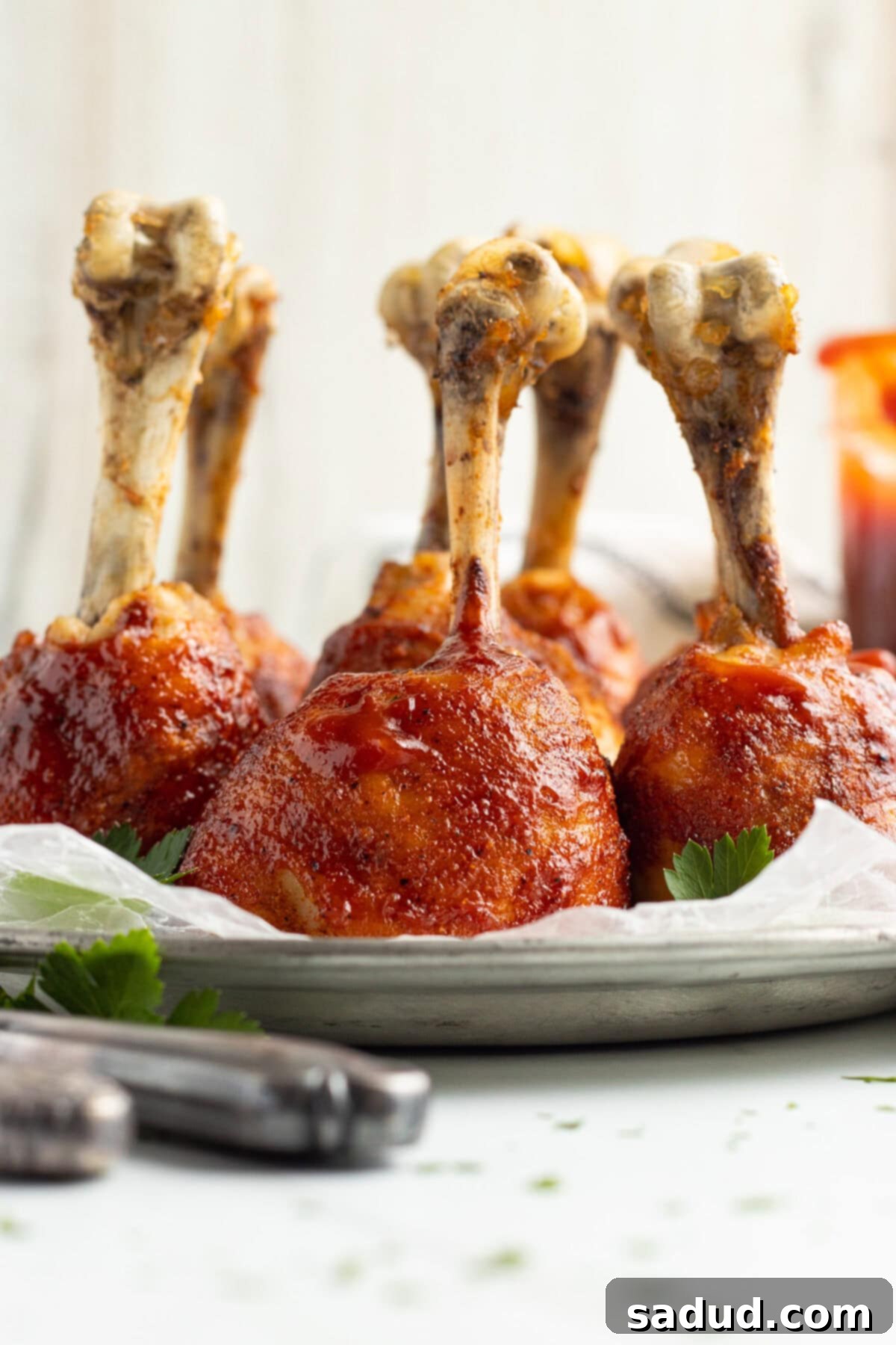 Side view of lollipop chicken legs coated in bbq sauce, standing upright on a baking sheet lined with parchment paper.