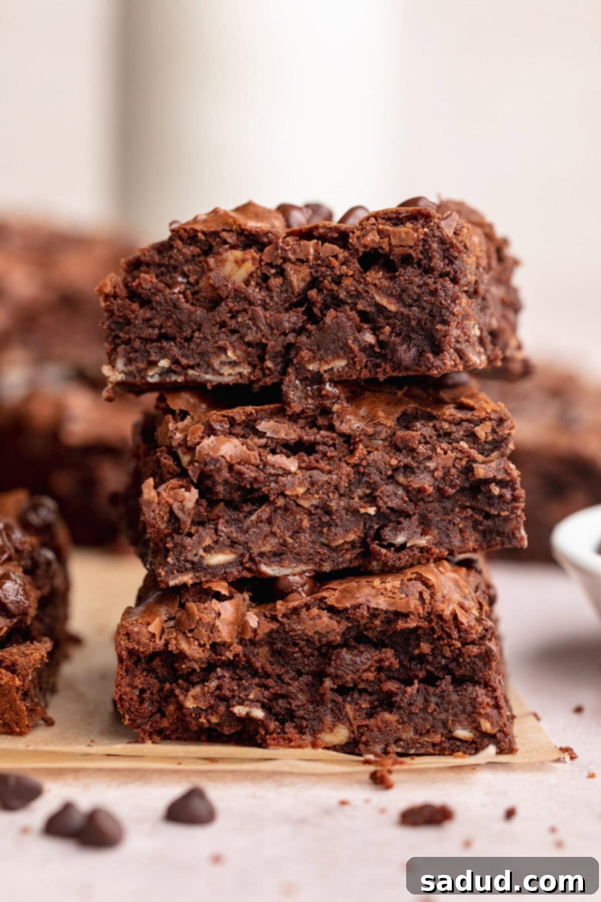 Three thick, rich, chocolatey lactation brownies stacked on top of each other, with chocolate chips and crumbs on the counter around them, suggesting a decadent, homemade treat.