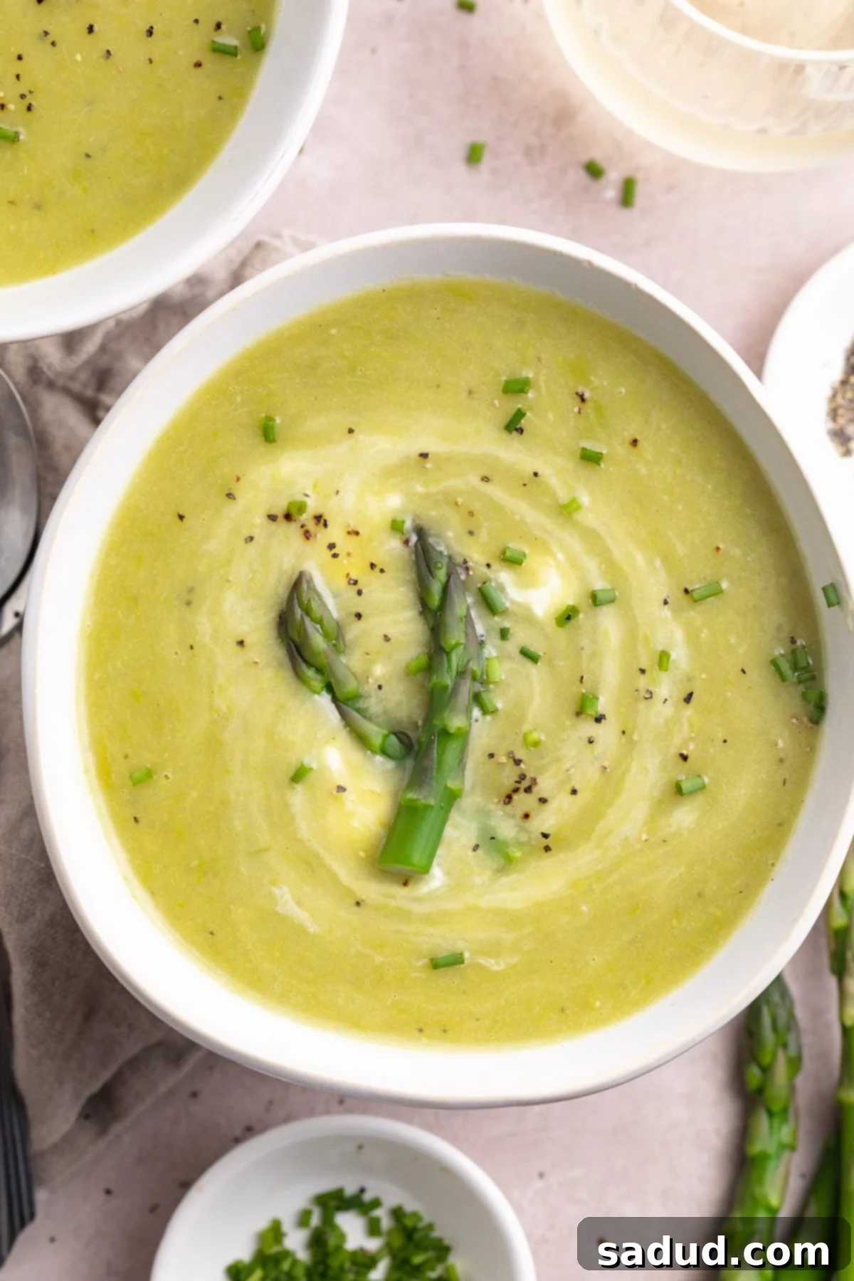 Top-down, overhead view of a white, misshapen round bowl holding pale green creamy potato soup garnished with asparagus stalks.