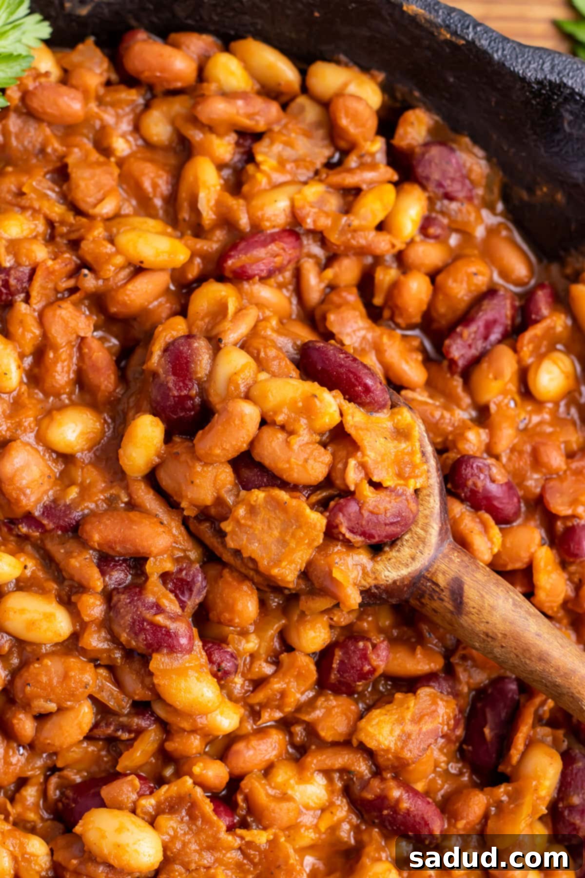 Close-up of perfectly smoked baked beans in a large cast-iron skillet, featuring visible bacon bits and a large wooden serving spoon.