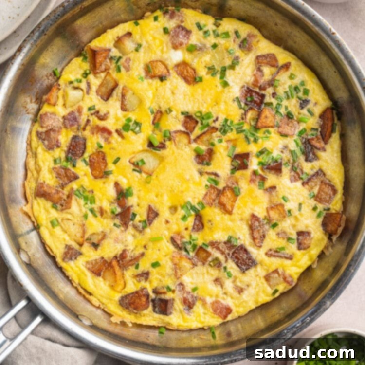 A large, golden-brown country omelette in a silver oven-safe skillet, garnished with fresh chives, ready to be sliced and served.