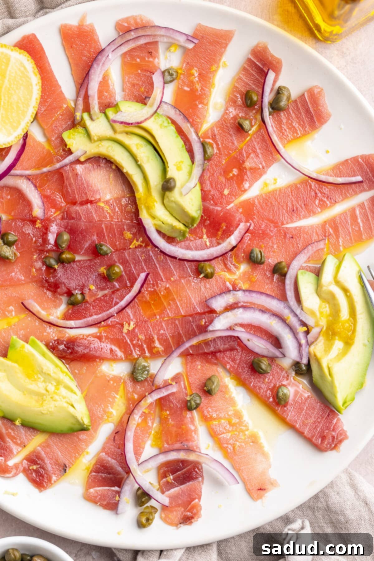 Tuna carpaccio arranged on a large white platter, topped with slices of avocado and red onion, with dots of capers. This image showcases the fresh, vibrant colors and elegant presentation of the dish.
