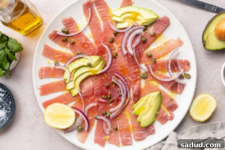 Tuna carpaccio arranged on a large white platter, topped with slices of avocado and red onion, with dots of capers.