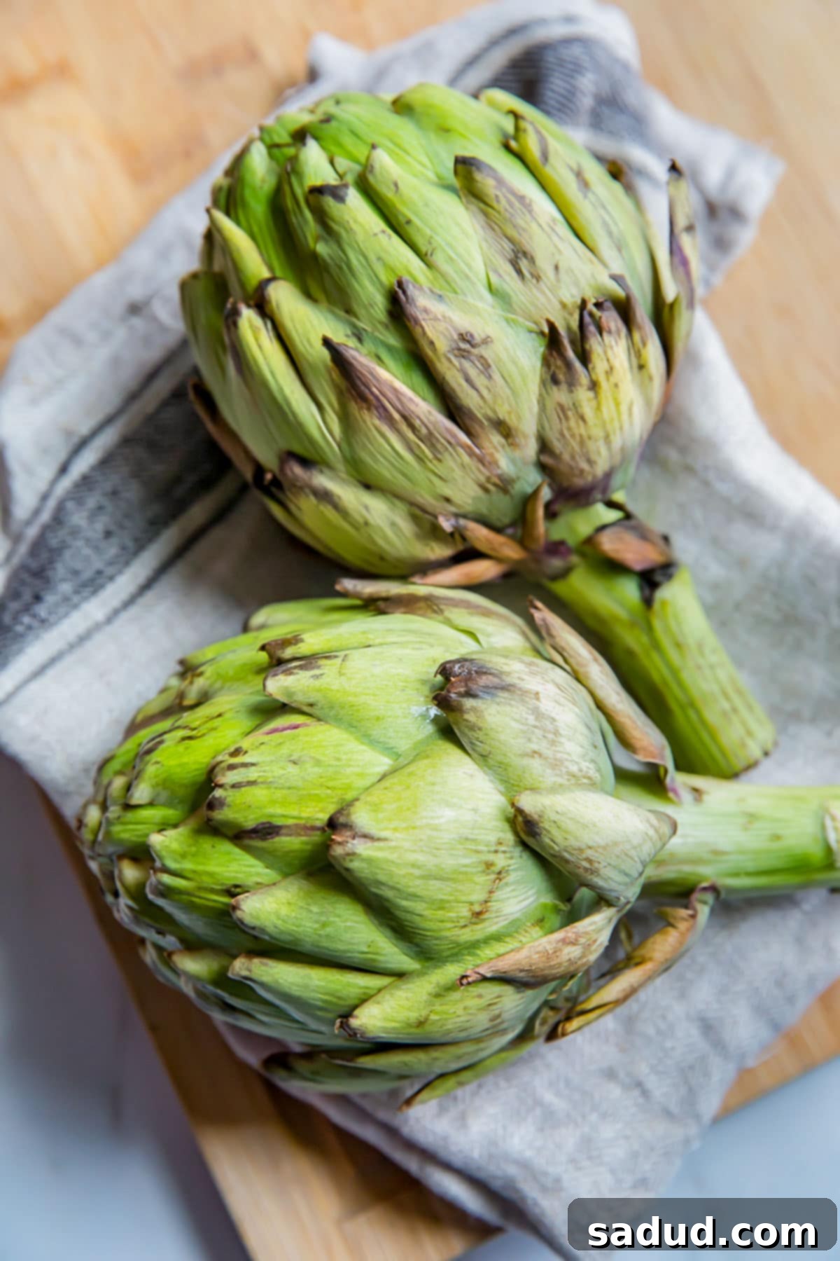 Two fresh, unboiled artichokes on a cutting board with a kitchen towel, ready for preparation.