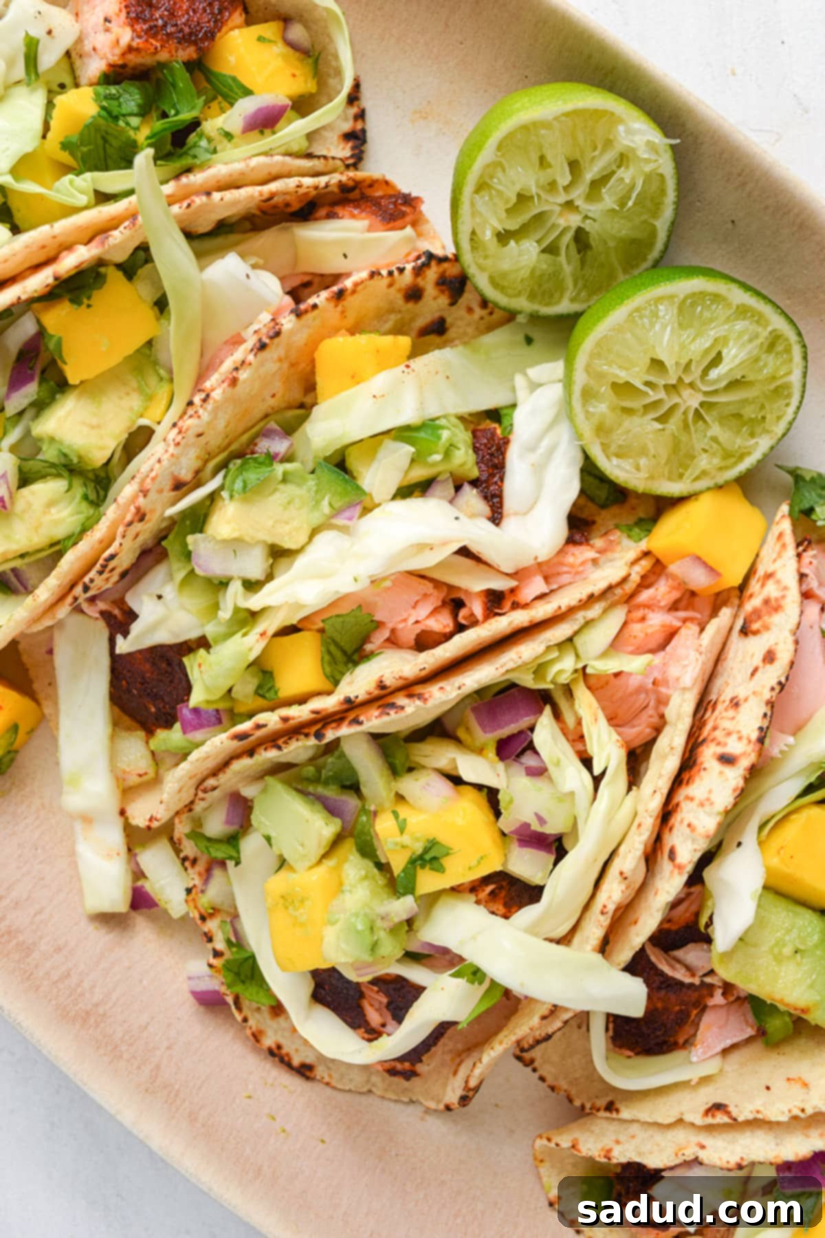 Four beautifully arranged salmon tacos, generously topped with fresh cabbage slaw and sweet mango salsa, presented on an elegant oval platter.