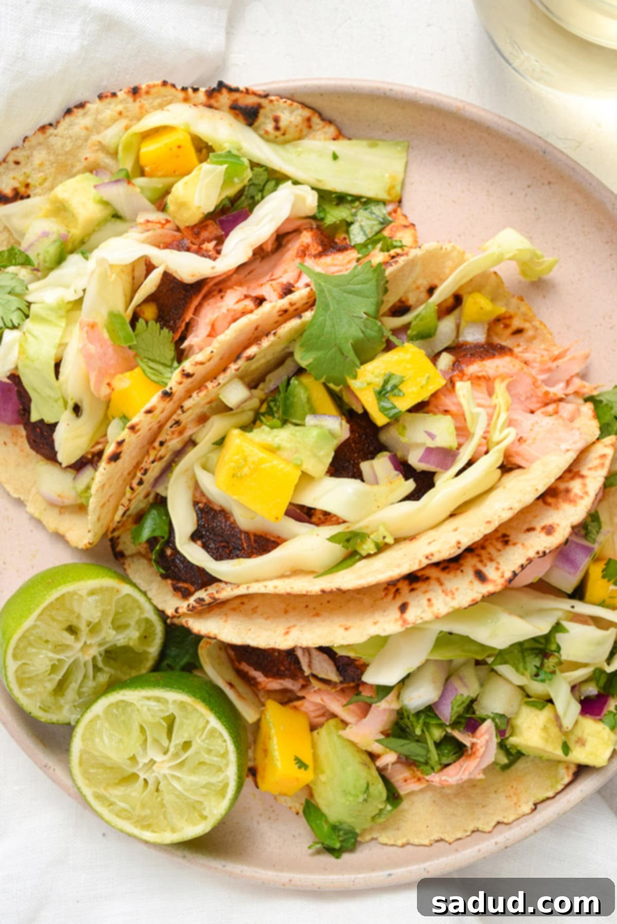 Three perfectly plated salmon tacos, adorned with luscious mango salsa and crisp fresh cabbage slaw, served on a stylish round plate.
