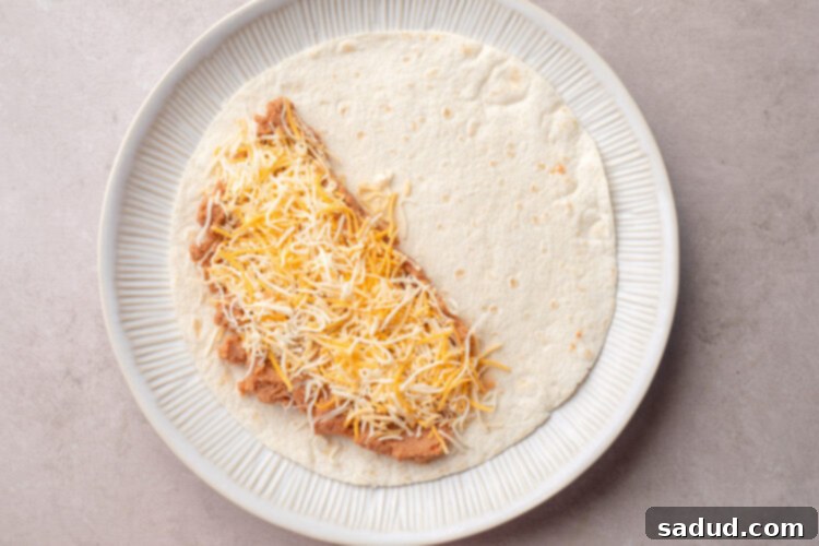 Refried beans and shredded cheese on one half of a 10-inch flour tortilla on a plate.