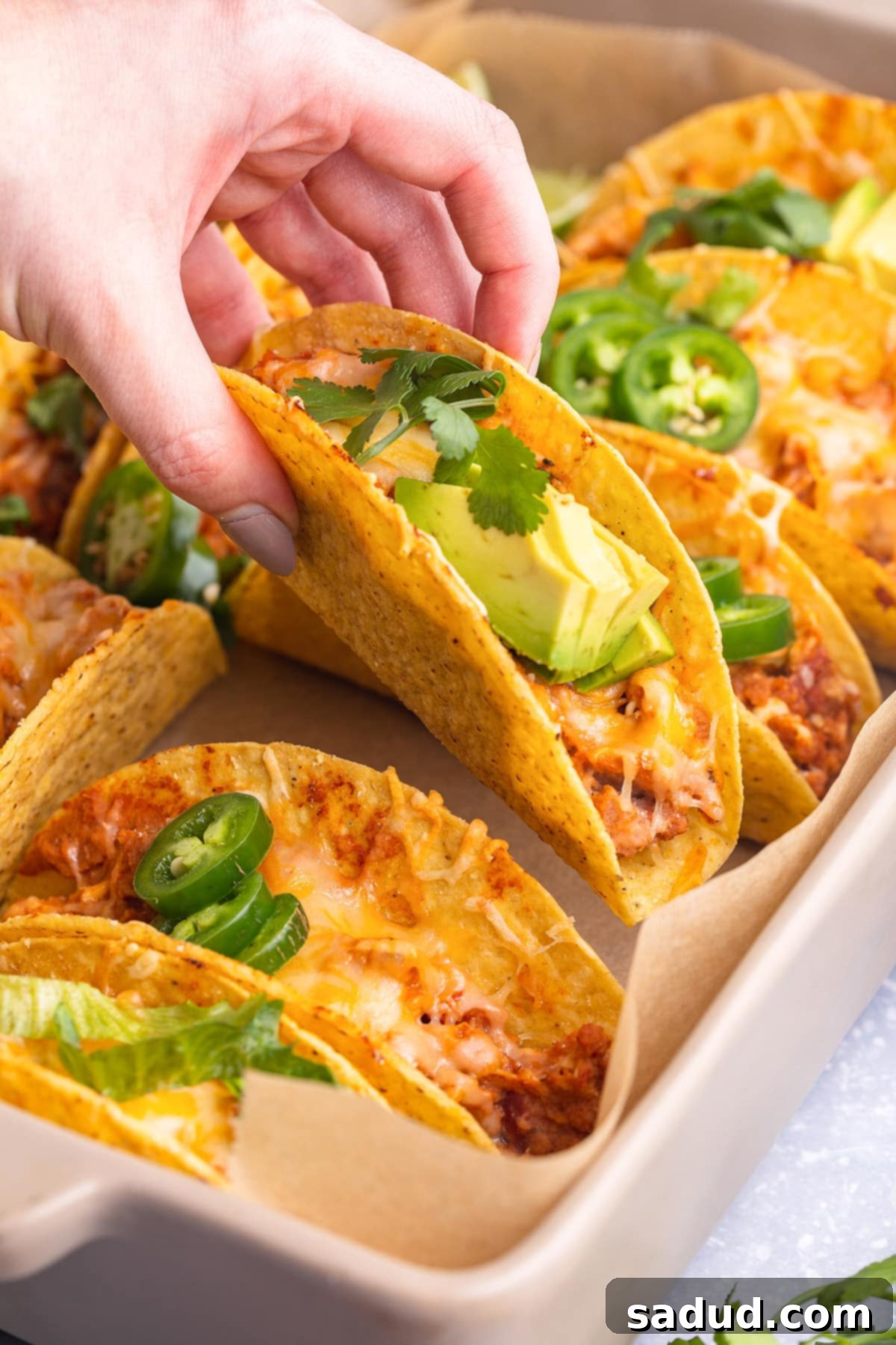A white woman's hand lifts a baked chicken taco in a crunchy shell out of a baking pan of other tacos.