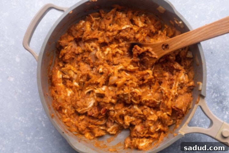 Shredded chicken, refried beans, salsa, taco seasoning, and onions cooking in a large skillet with a wooden spoon.