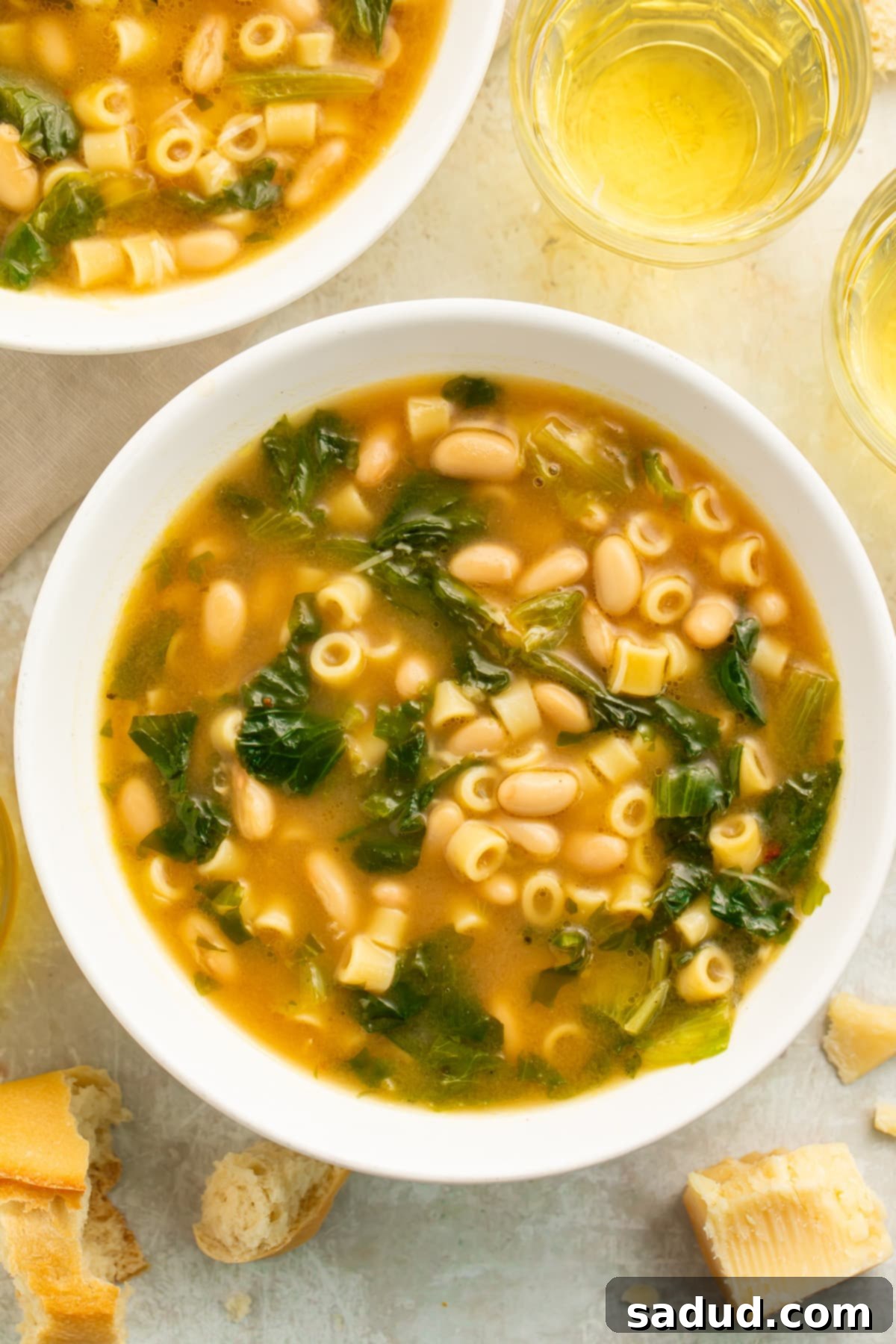 Zoomed-out, overhead view of a white soup bowl filled with escarole soup, made up of short tube noodles, beans, and greens.