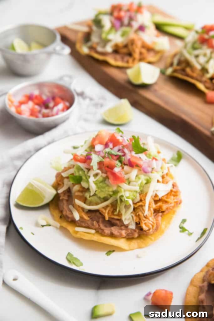 A chicken tostada on a white plate with lettuce, cheese, guacamole and salsa