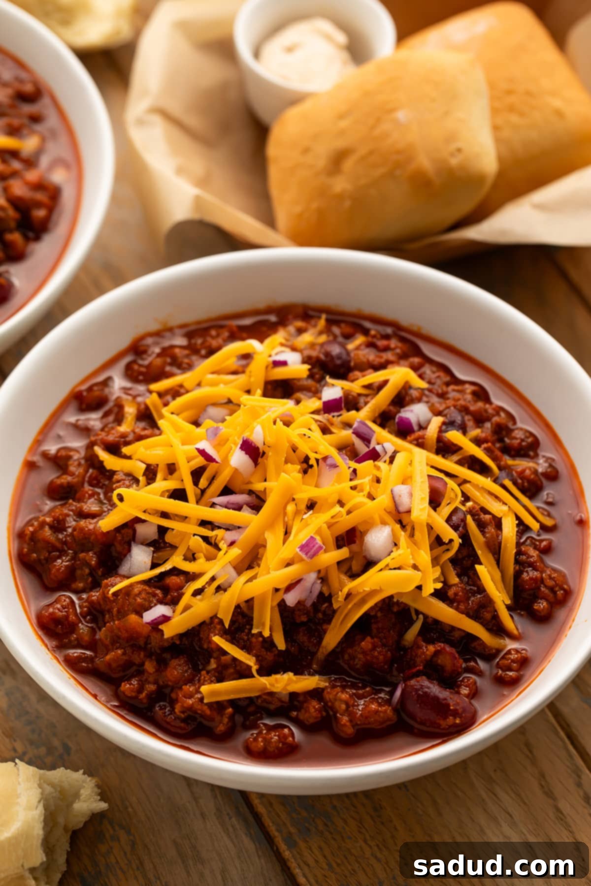 A perfectly angled view of a white ceramic bowl filled with dark red Texas Roadhouse chili, generously topped with vibrant shredded cheddar cheese and finely diced red onions. In the background, a rustic basket holds freshly baked, warm yeast rolls, inviting you to dip them into the savory chili. The scene captures the ultimate comfort food experience.