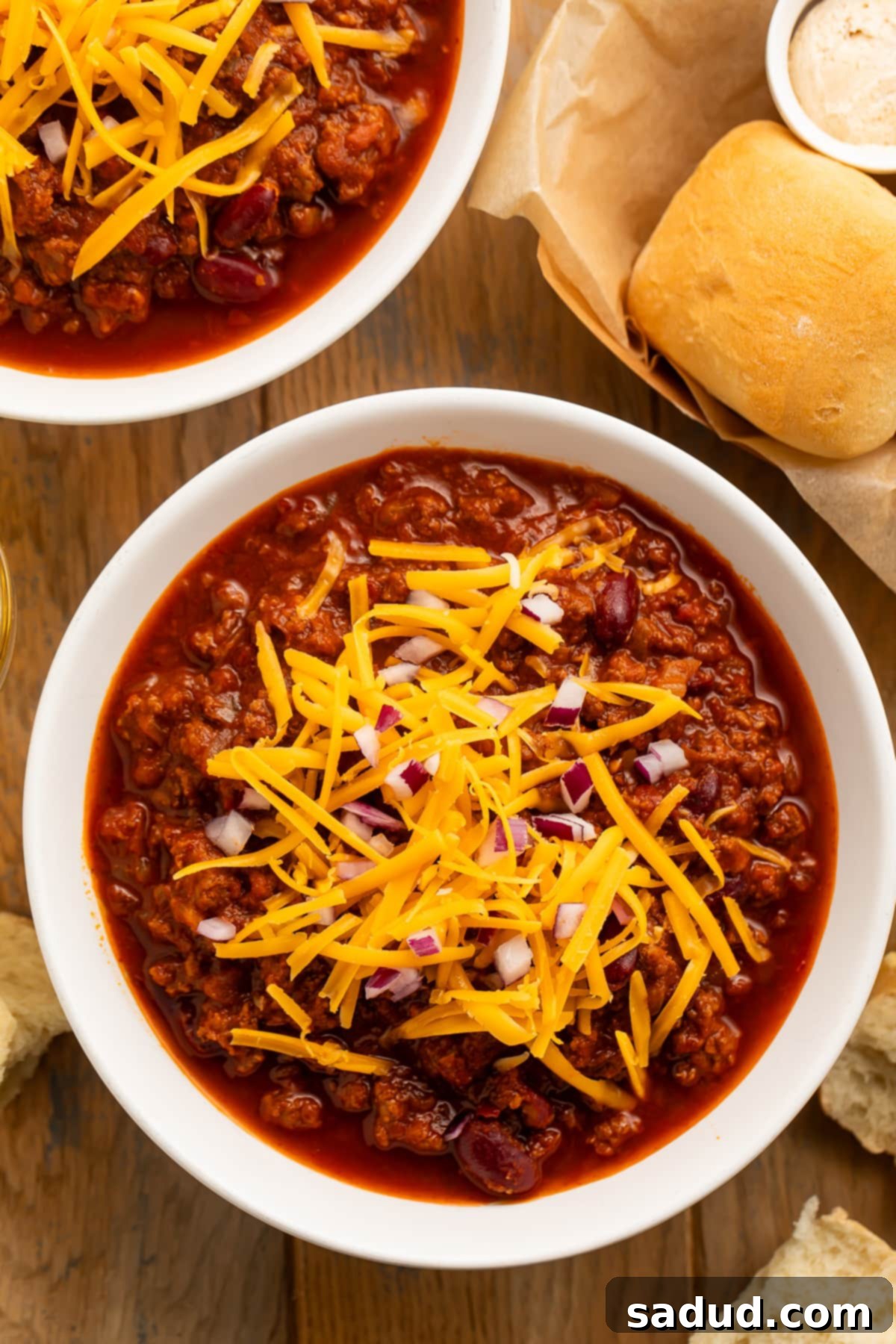 An inviting overhead view of a bowl of dark red, hearty Texas Roadhouse chili. It's generously adorned with bright orange shreds of fresh cheddar cheese and perfectly diced red onions, creating a beautiful contrast on a dark tabletop, ready to be enjoyed as a comforting meal.