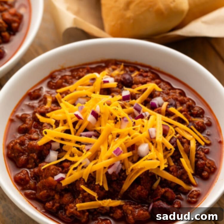 Angled view of a white bowl holding deep red chili topped with shredded cheddar cheese and diced red onions in front of a basket of rolls.