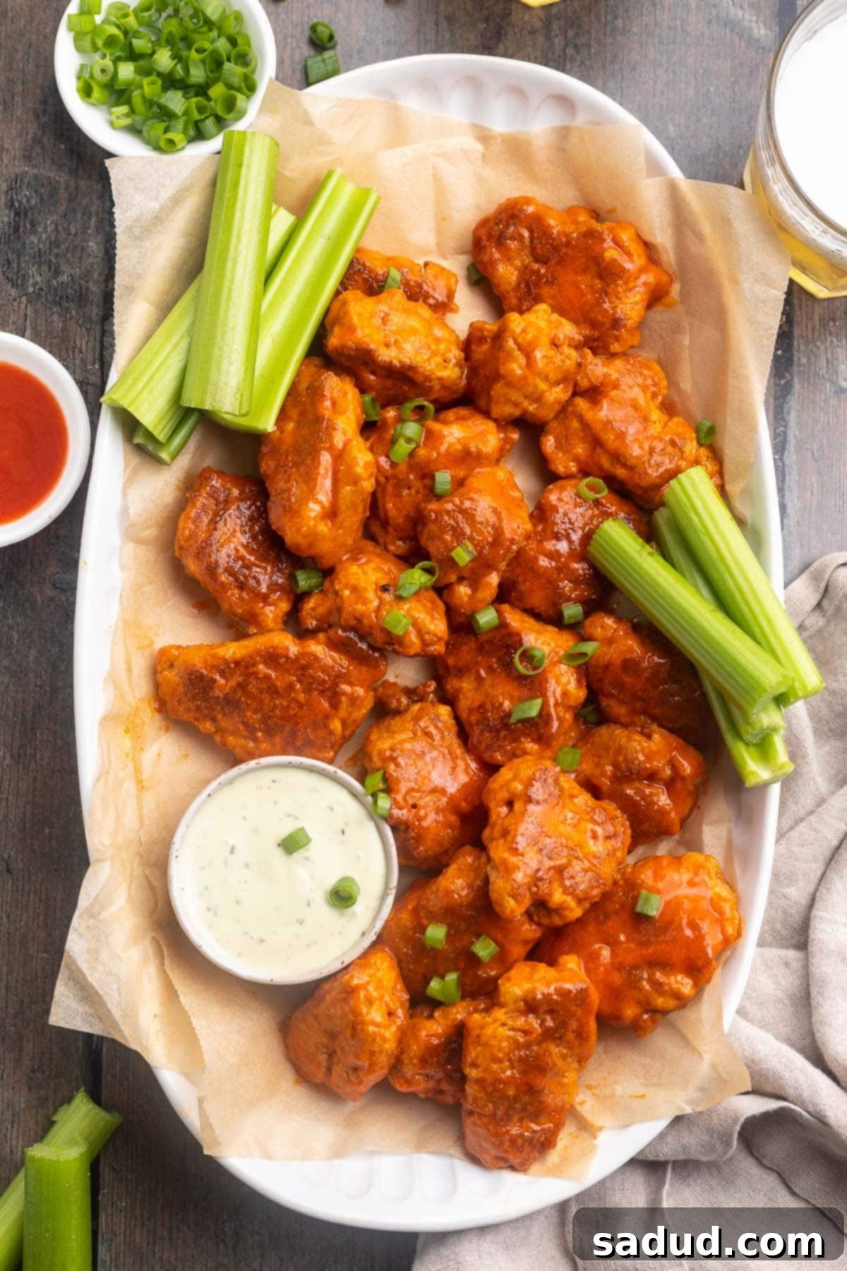 A vibrant platter of golden-brown buffalo sauced seitan wings, garnished with fresh parsley and served with a side of creamy vegan ranch dressing and crisp celery sticks for dipping.