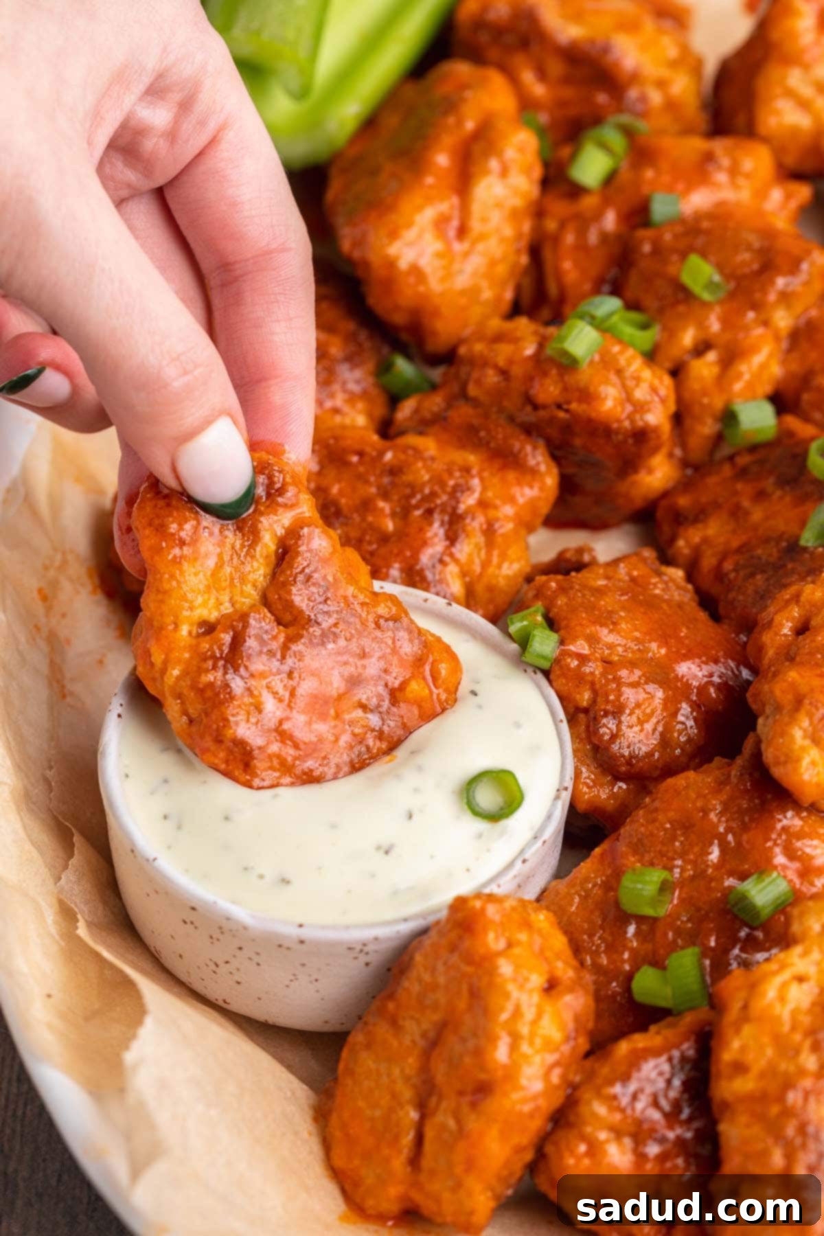 A hand gently dipping a crispy vegan seitan buffalo wing into a small white bowl of creamy vegan ranch dressing, showcasing its delicious texture.
