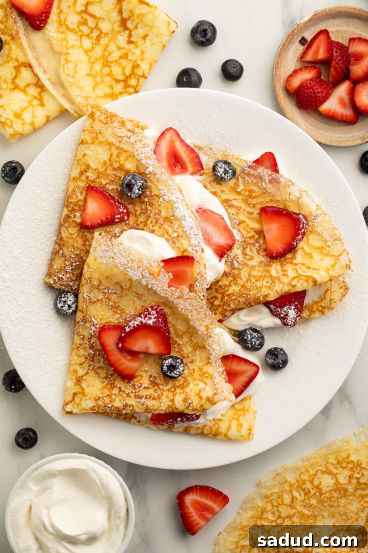 A pile of gluten free crepes filled with whipped cream and topped with sliced strawberries, fresh blueberries, and a light dusting of powdered sugar.