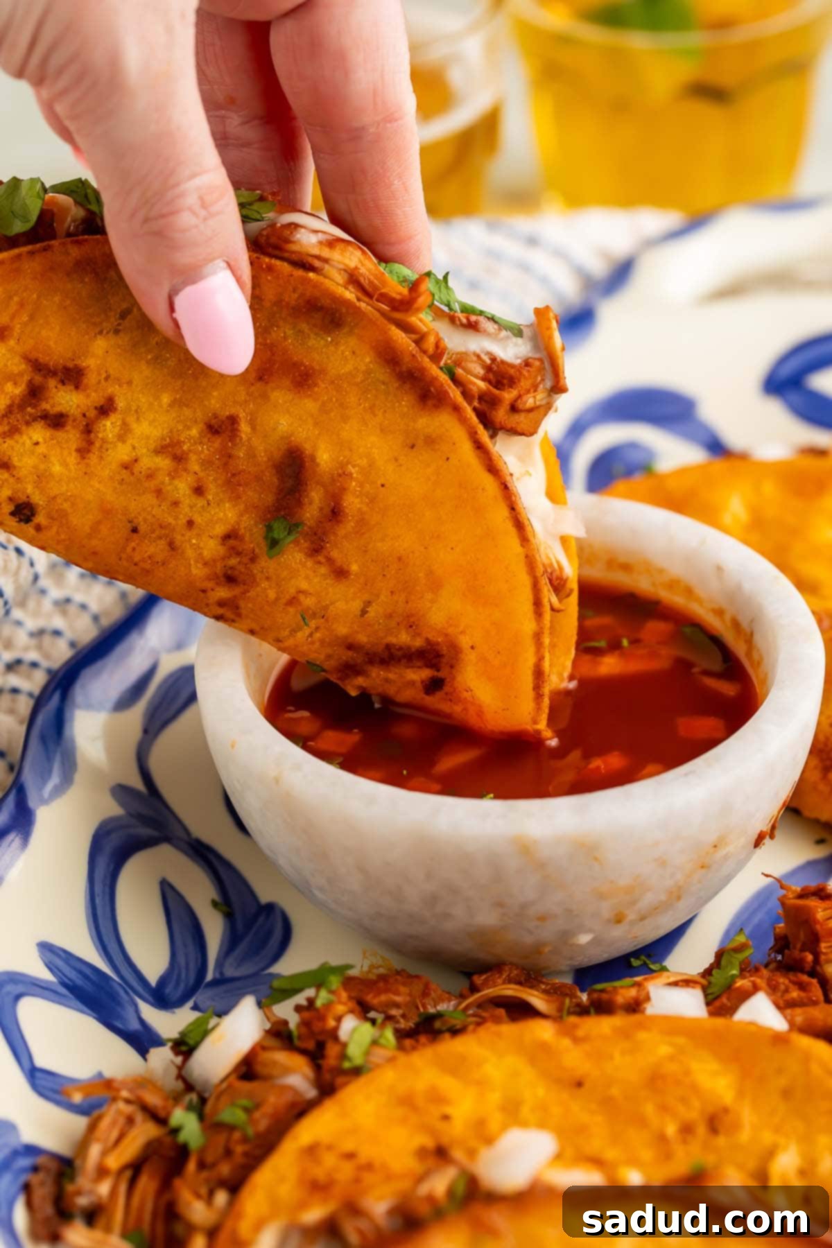 A crispy vegan birria taco filled with spiced jackfruit and melty cheese being dipped into a rich, red chile consommé, showcasing the immersive eating experience.