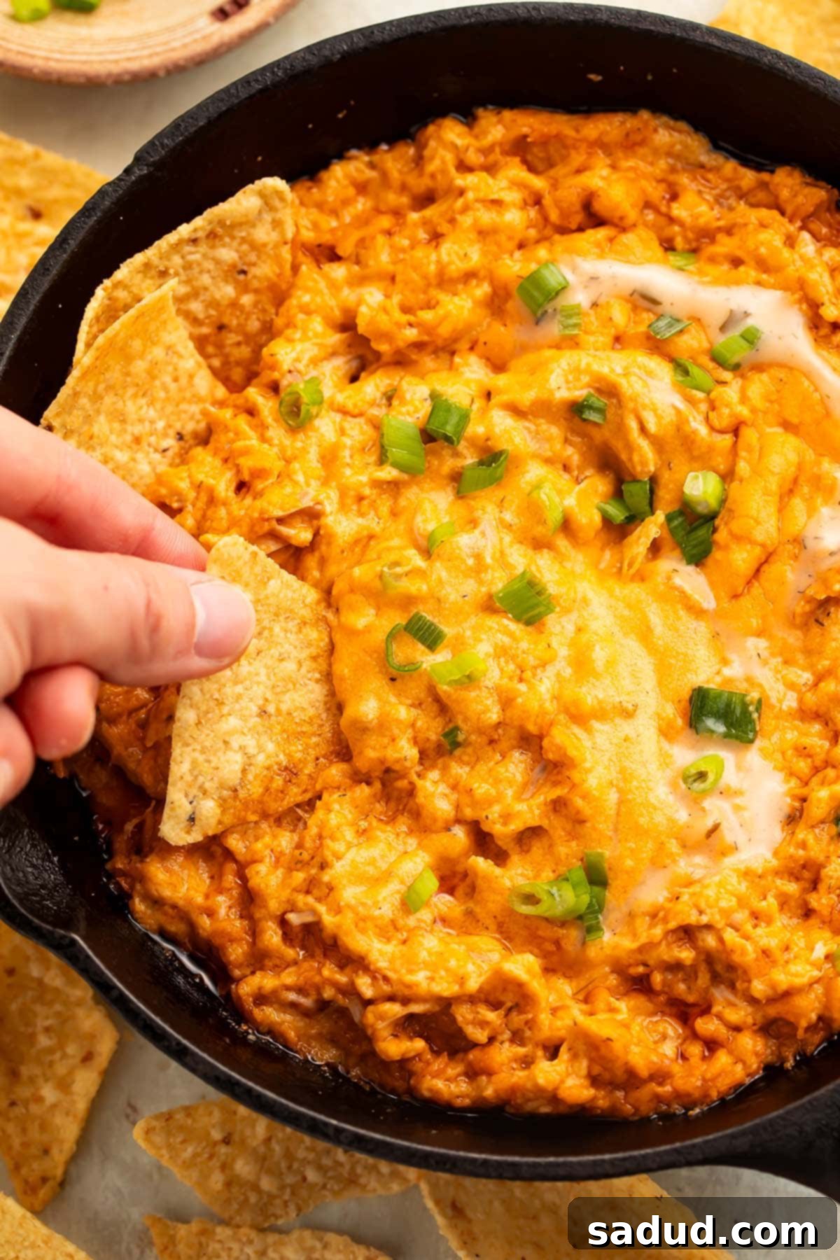 A woman dips a tortilla chip into a large cast-iron skillet holding creamy vegan buffalo chicken dip, showing its perfect texture and rich color.