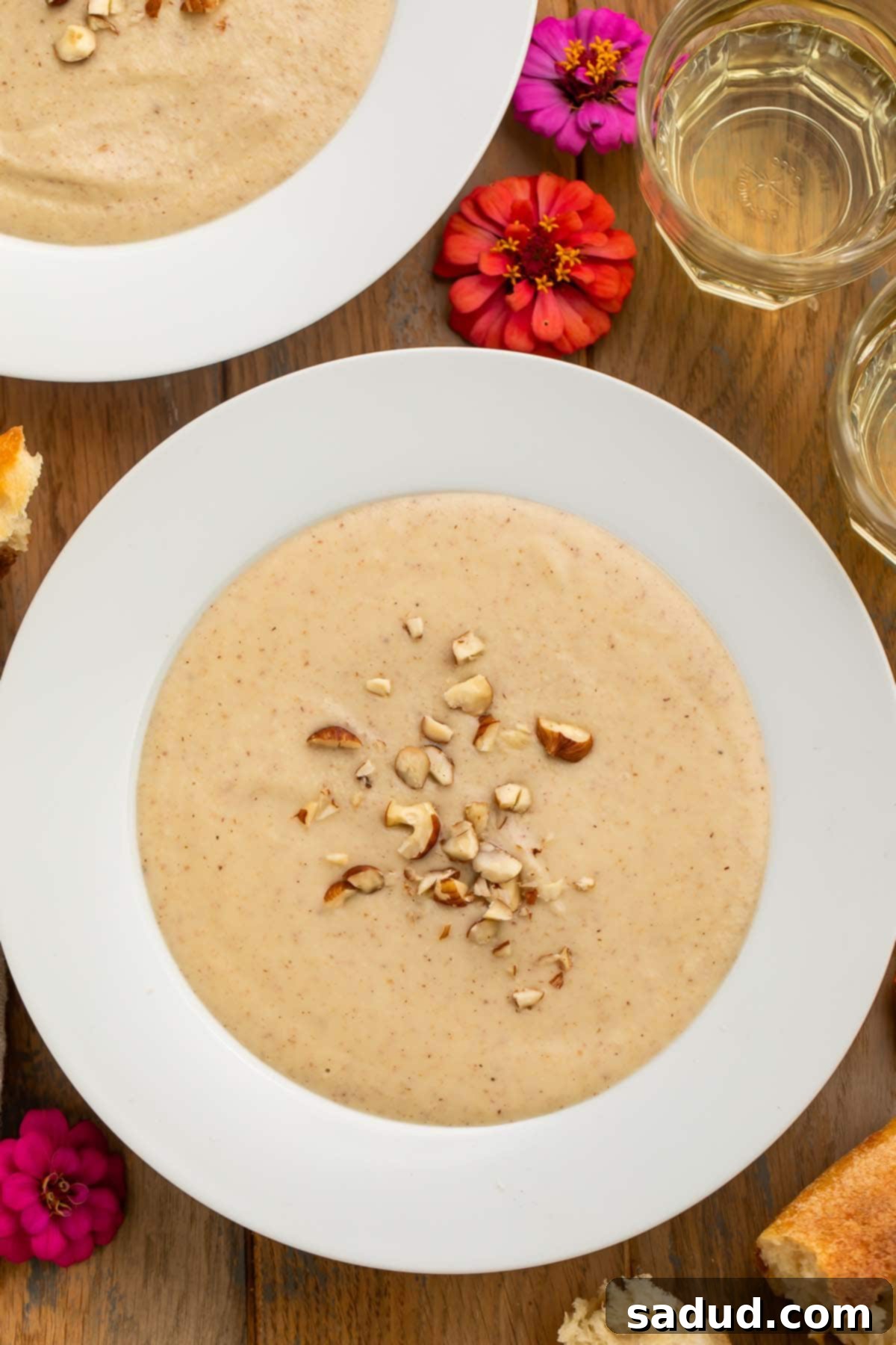 Roasted Parsnip and Hazelnut Soup 2 A large white bowl of creamy, pale hazelnut soup, garnished with chopped hazelnuts and fresh thyme, rests on a rustic wooden table adorned with delicate pink flowers, evoking a cozy and inviting scene.