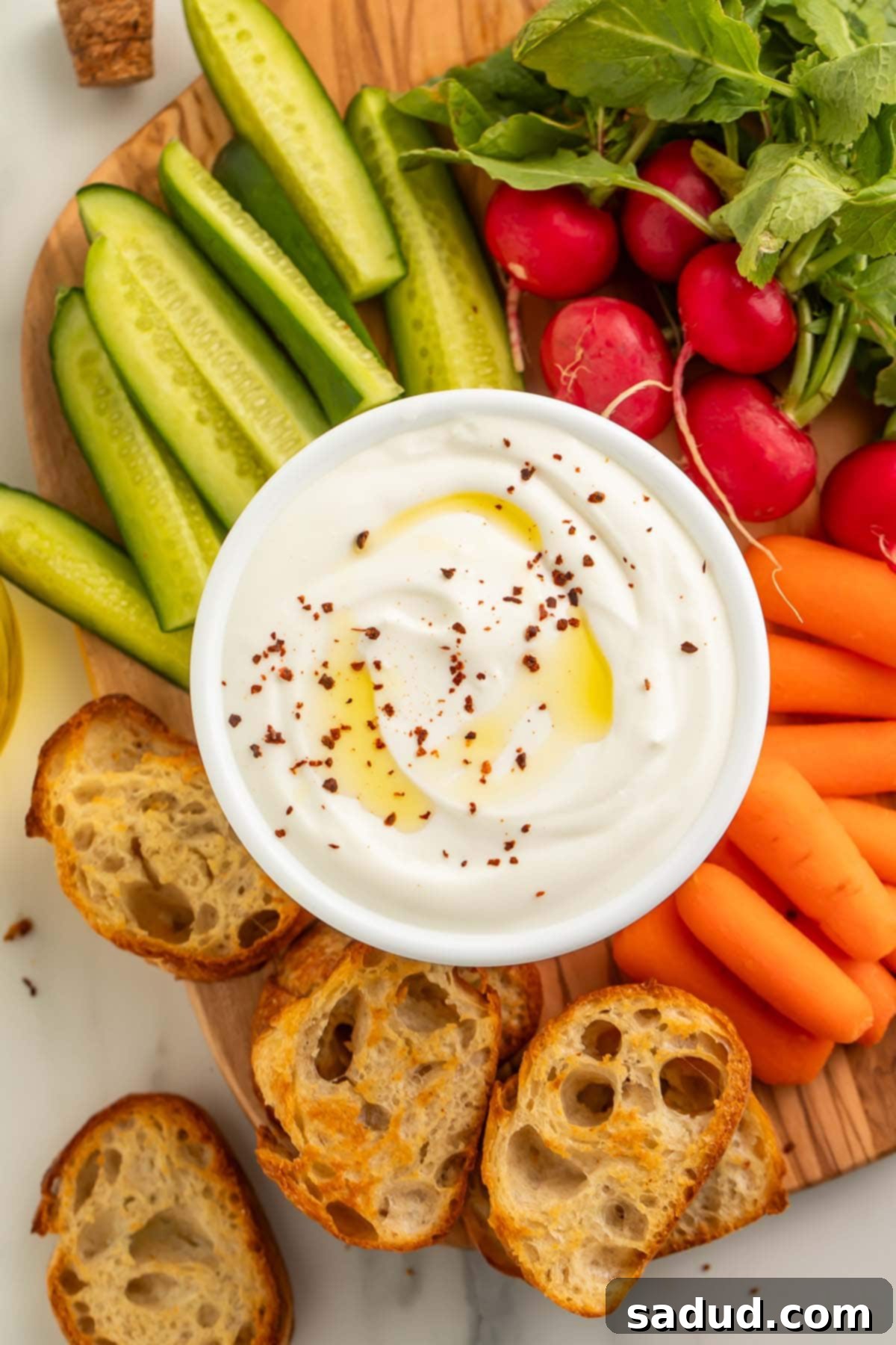 A bowl of savory whipped cottage cheese resting on a wooden board, surrounded by crostini, pickles, carrot sticks, and radishes.