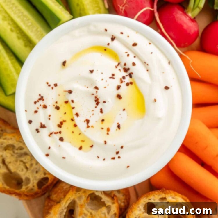 A bowl of savory whipped cottage cheese resting on a wooden board, surrounded by crostini, pickles, carrot sticks, and radishes.