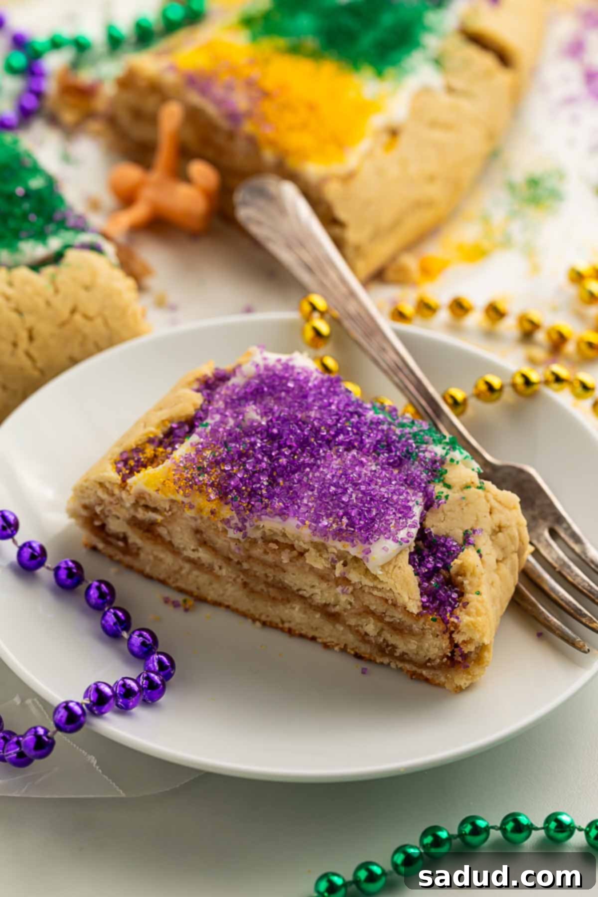 A delectable slice of gluten-free King Cake, topped with vibrant purple sanding sugar, elegantly presented on a white plate with a silver fork, and surrounded by colorful purple, gold, and green Mardi Gras bead necklaces.