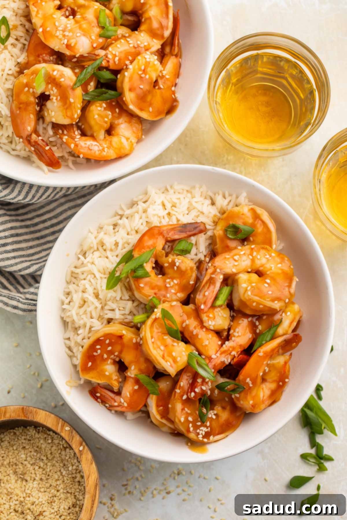 Bowls of teriyaki shrimp and cooked white rice on a table surrounded by green onions and a small bowl of sesame seeds, illustrating a complete meal setup.