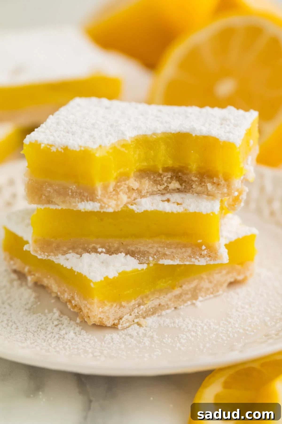 A stack of 3 beautifully golden vegan lemon bars, each generously topped with powdered sugar, resting on a pristine white plate, showcasing their vibrant yellow filling.