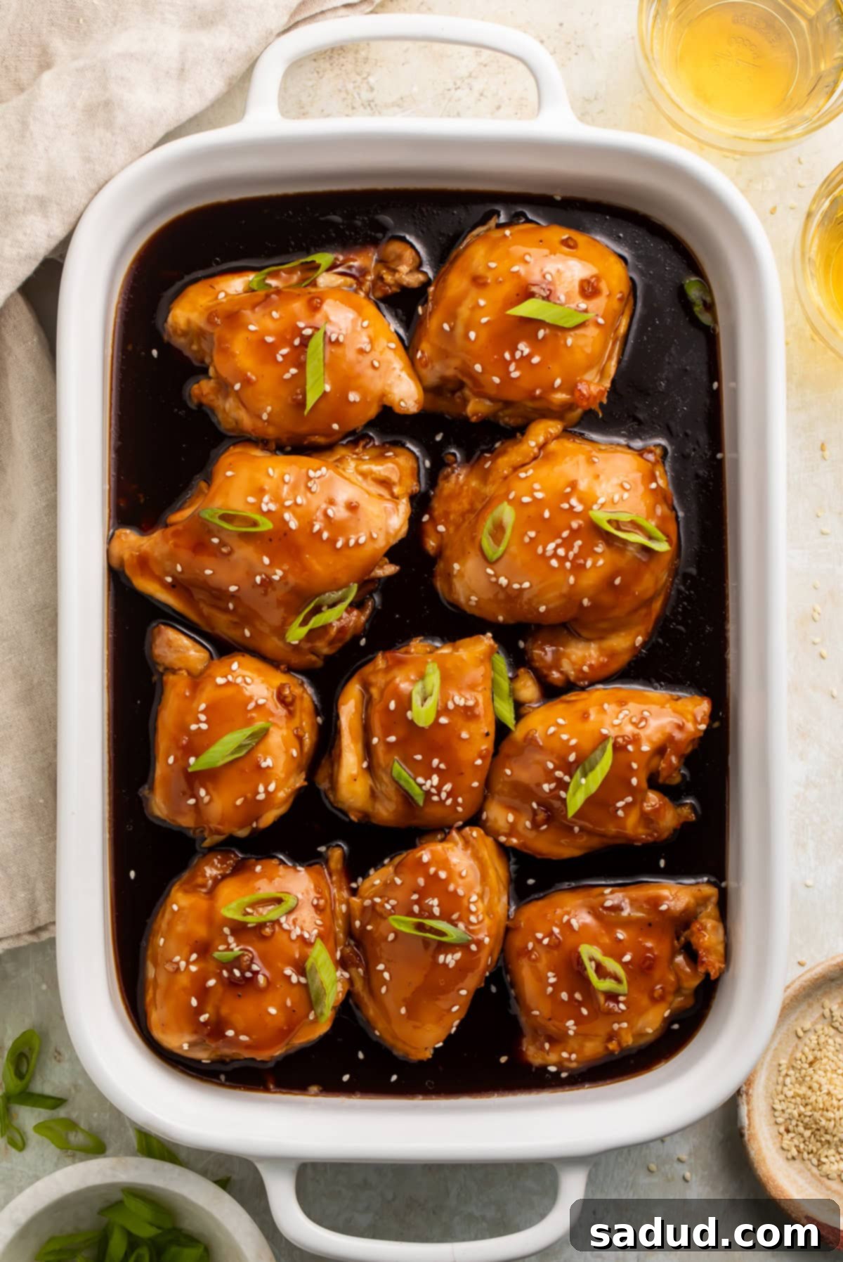 White baking dish with teriyaki chicken thighs and sauce, topped with sliced green onions and sesame seeds. 