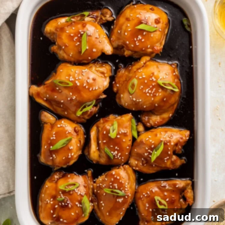 White baking dish with teriyaki chicken thighs and sauce, topped with sliced green onions and sesame seeds.