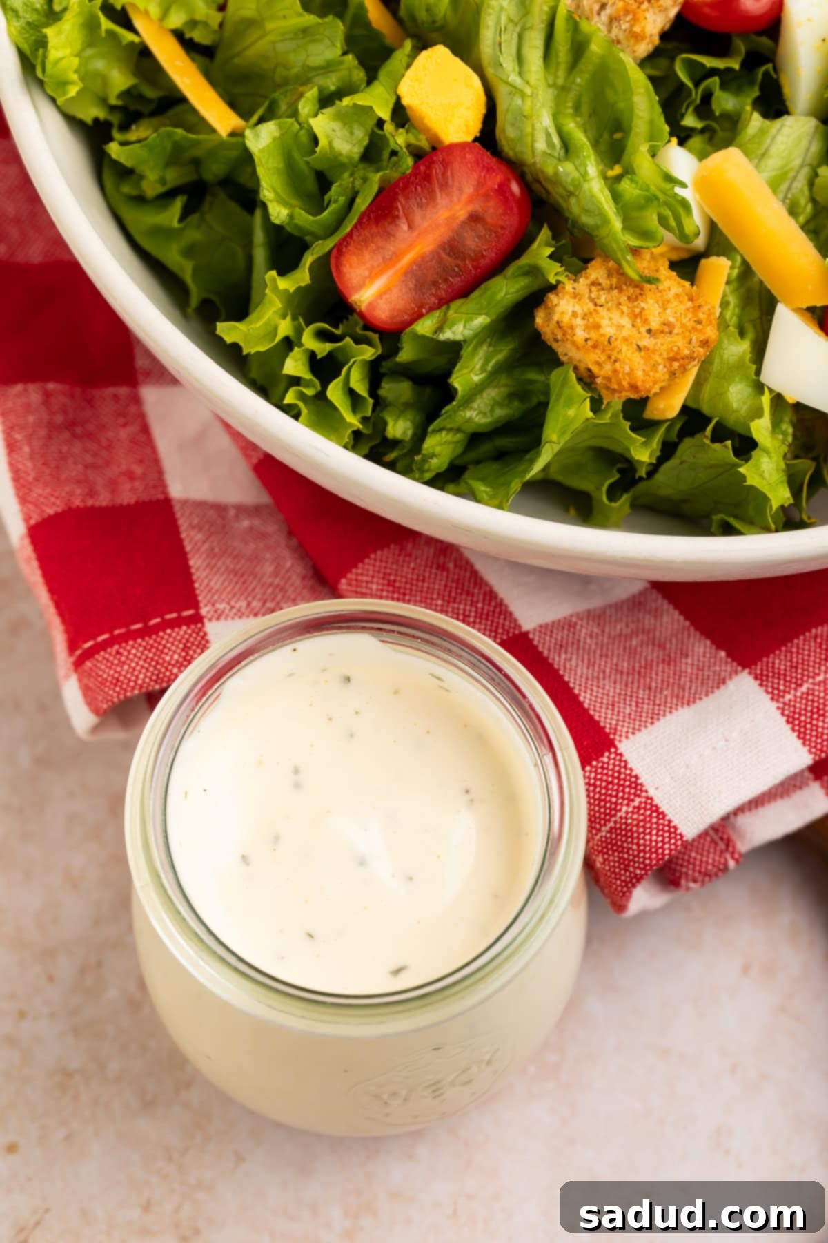 Top down view of a creamy ranch dressing in a glass jar next to a salad sitting on a gingham napkin. 