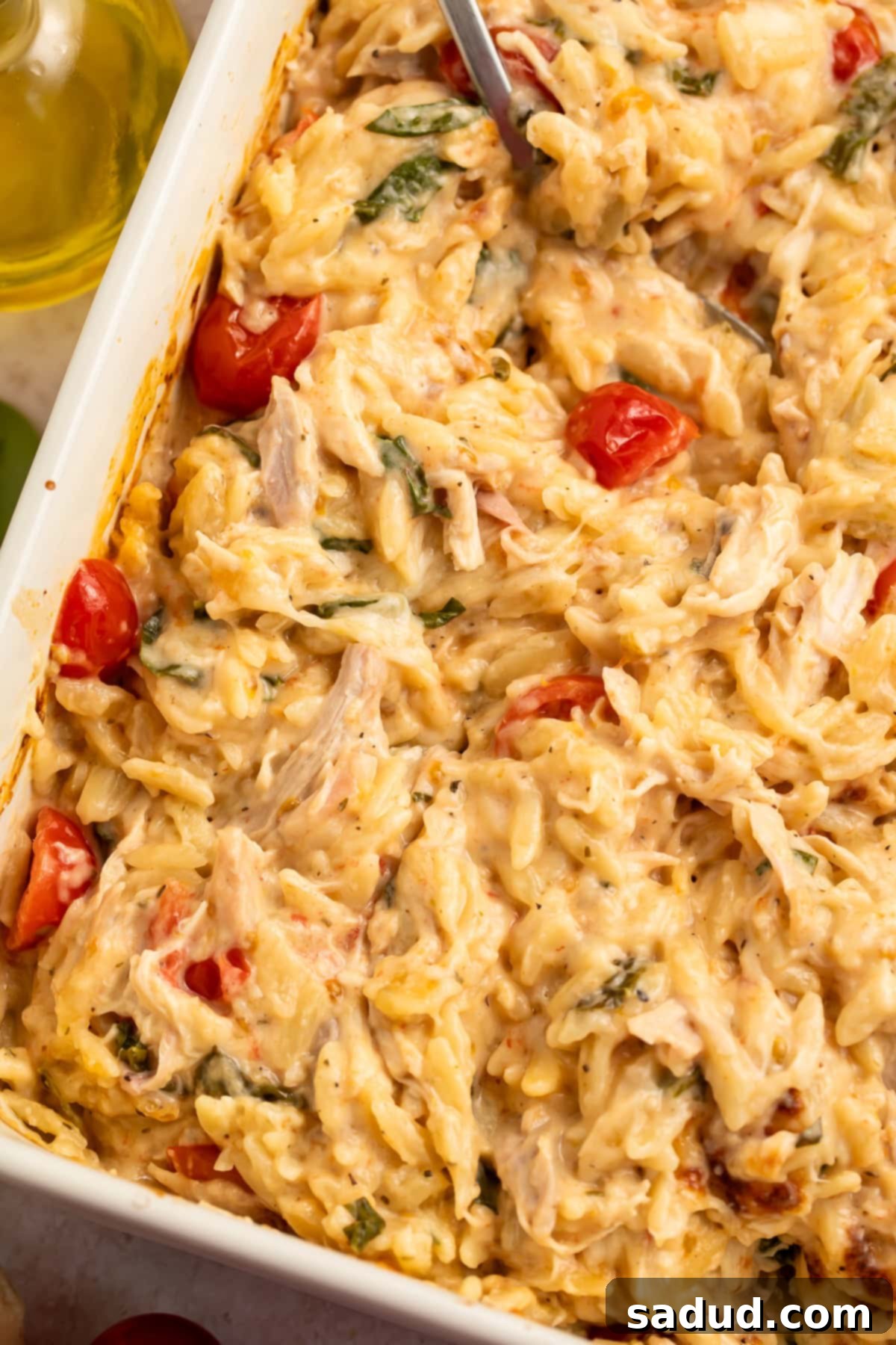 Golden Baked Chicken Orzo 2 Close up of a stirred chicken and orzo bake in a white baking dish, showing creamy pasta, chicken pieces, spinach, and melted cheese.