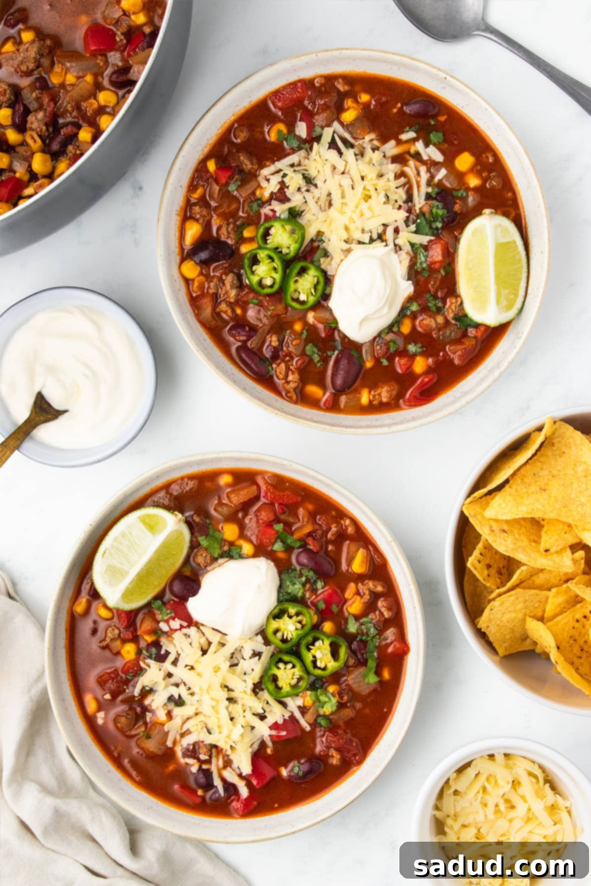 Two white bowls of chili surrounded bowls with fixings of sour cream and tortilla chips.