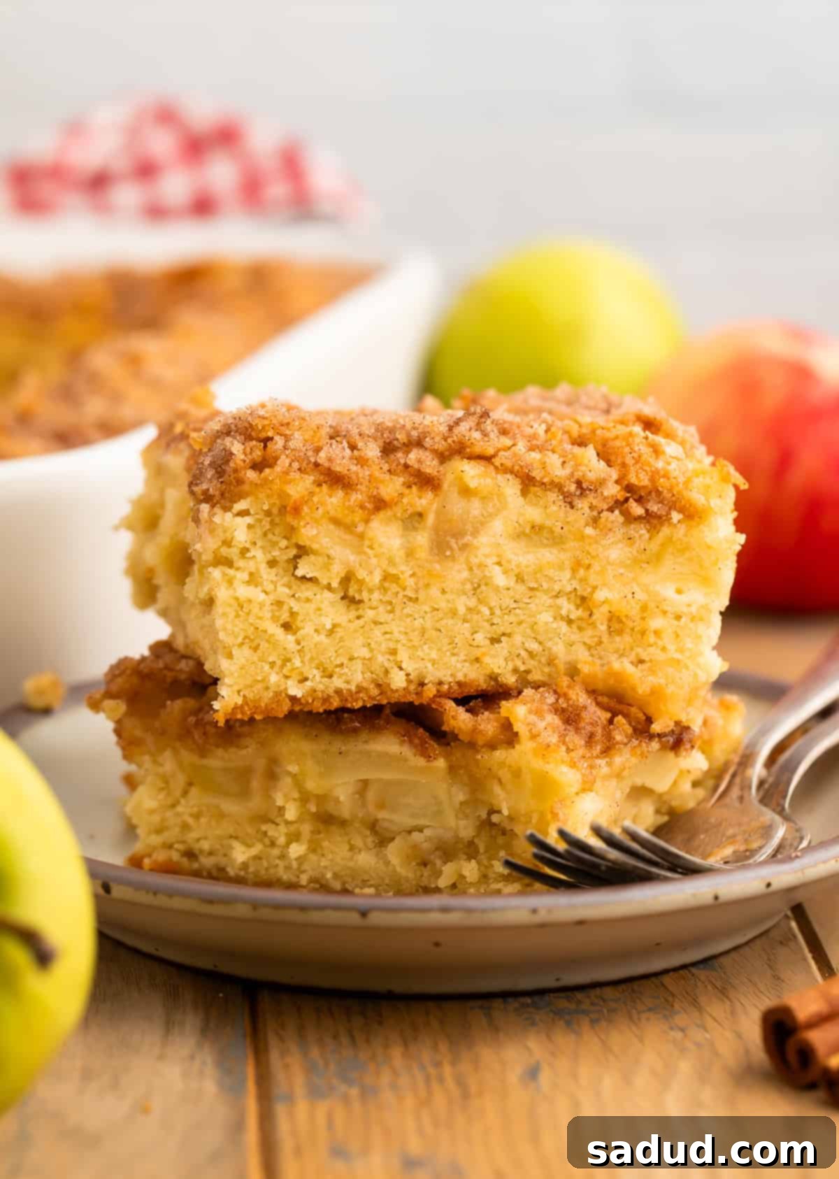 A perfectly sliced piece of gluten-free apple cake, showing the tender apple chunks and a golden cinnamon-sugar topping, rests on a grey plate. In the background, the full cake pan and fresh apples hint at the baking process and natural ingredients.