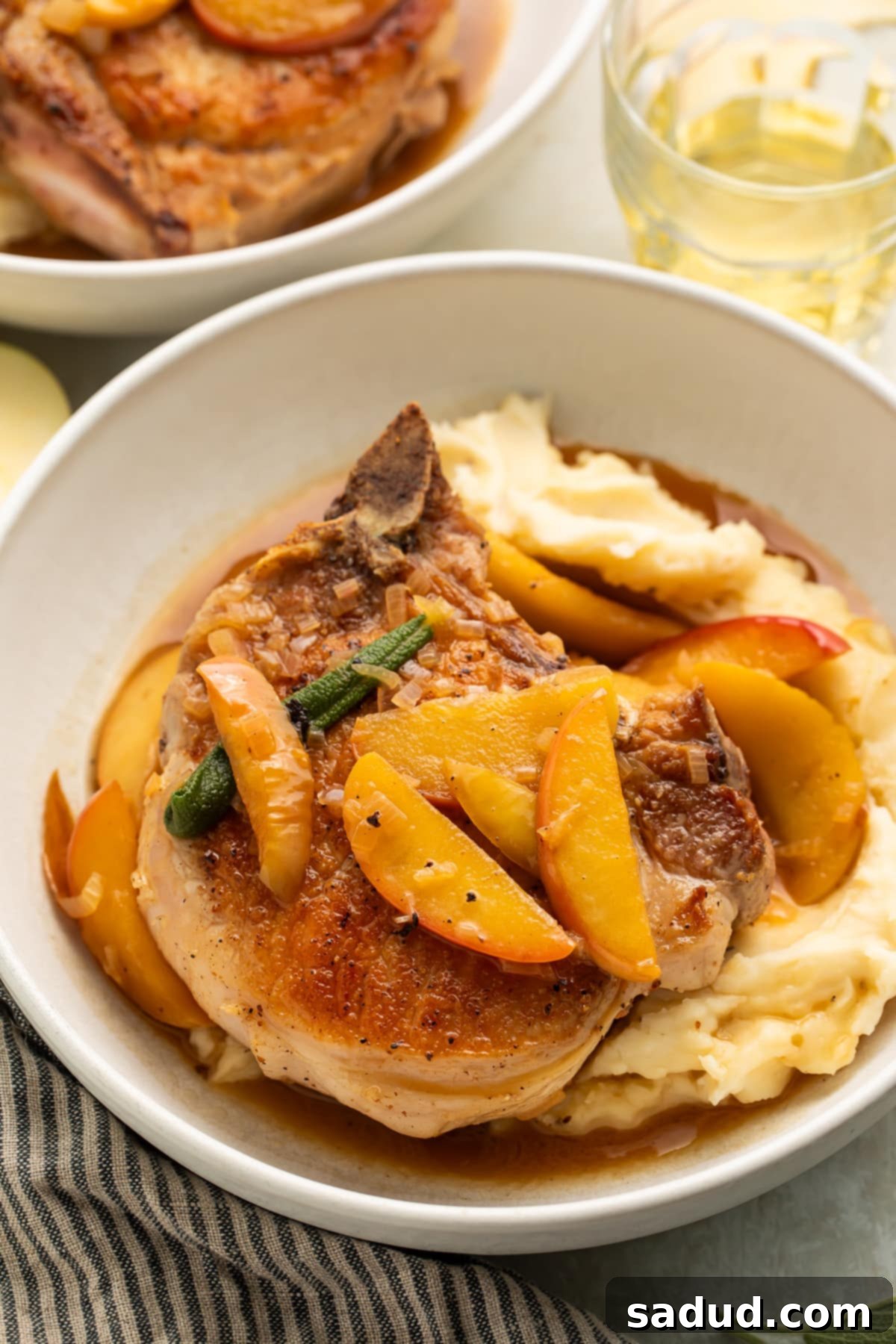 Seared pork chops with apples and a sage butter sauce over mashed potatoes in a large white bowl.