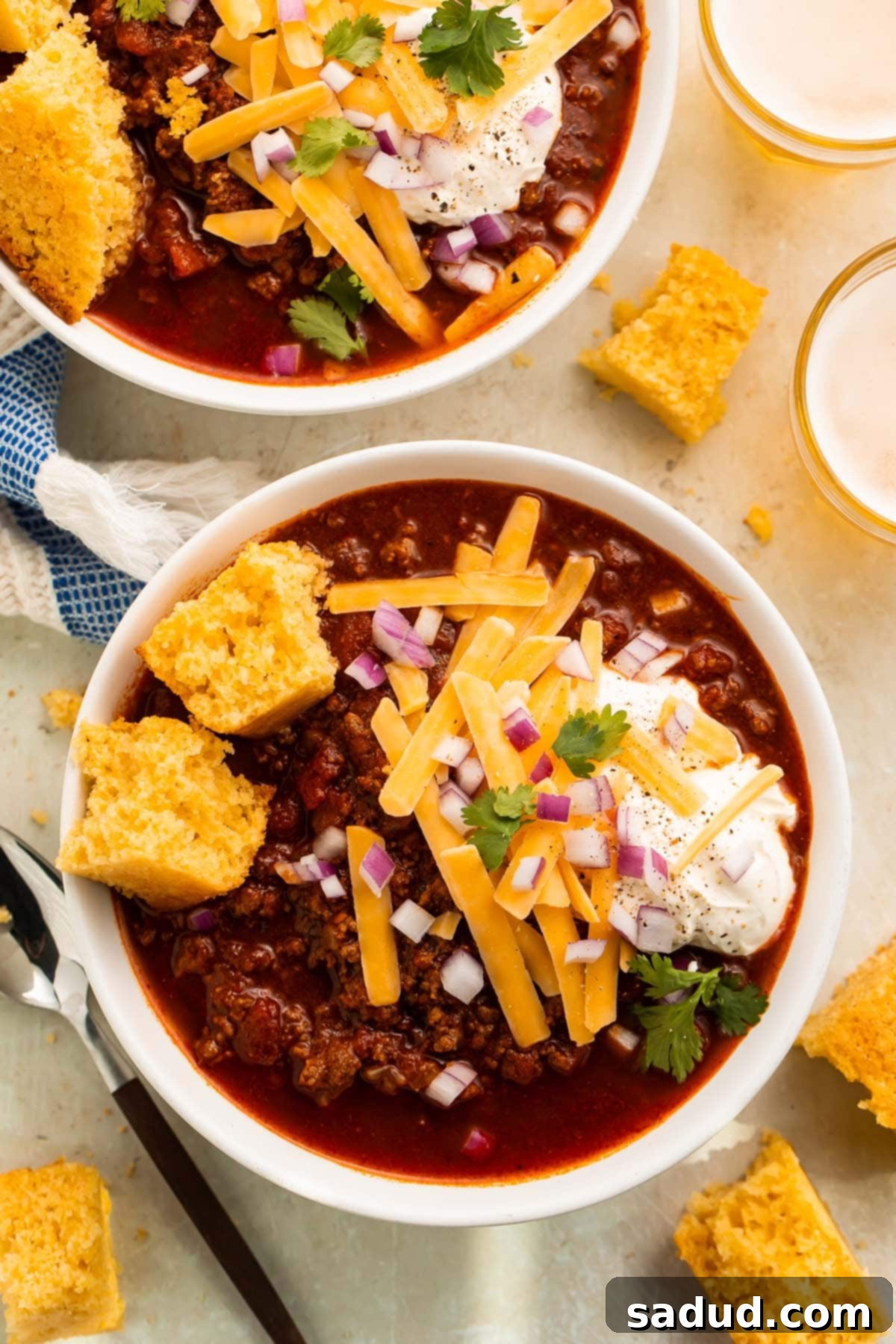 On a table: two white bowls containing deep red low carb chili topped with a large dollop of sour cream, thick shreds of yellow cheddar cheese, diced purple onion, and chopped parsley.