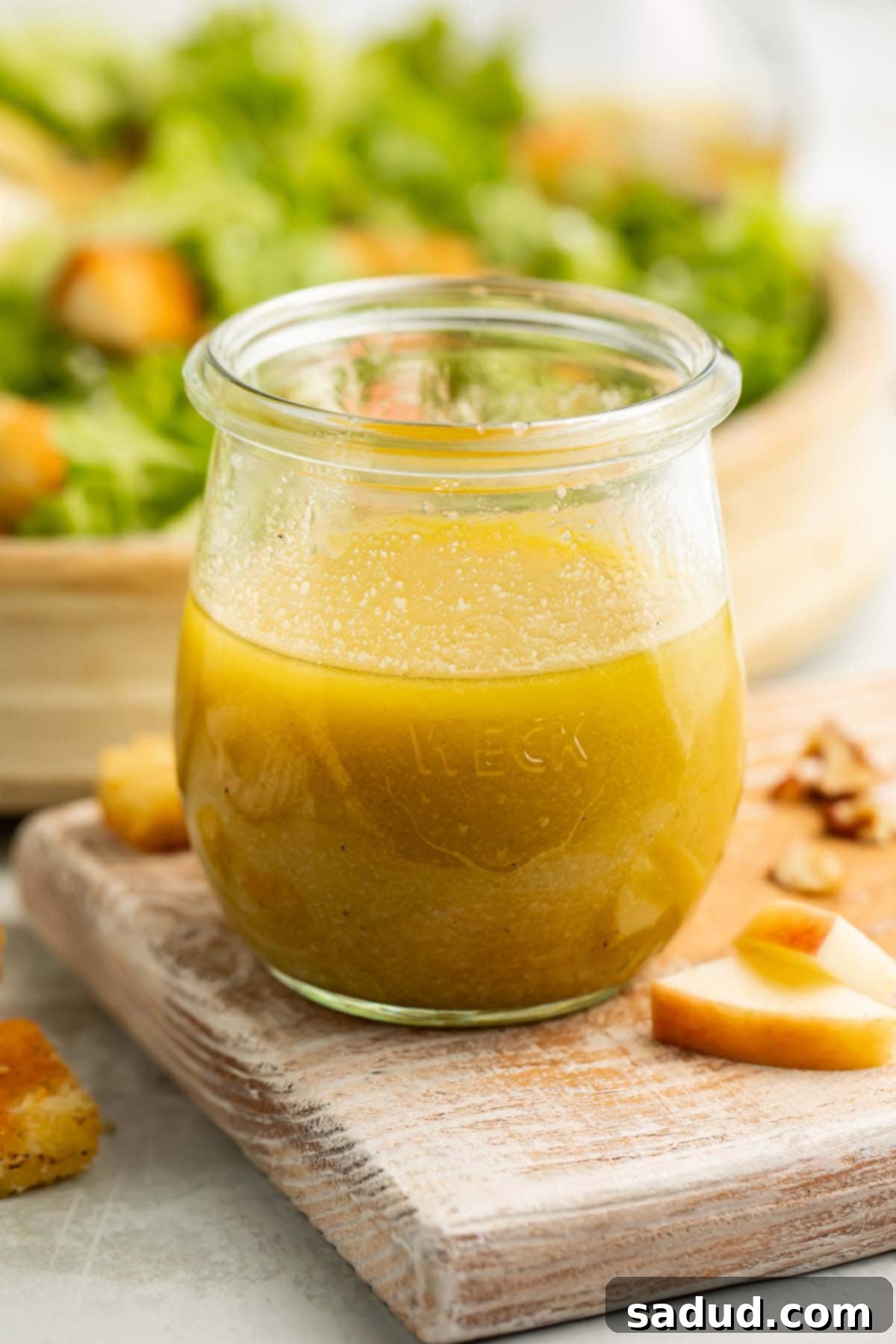 A glass jar of vibrant green apple cider vinaigrette rests on a rustic cutting board, surrounded by fresh apple slices and a lush green salad, evoking a sense of fresh, healthy, and homemade goodness.