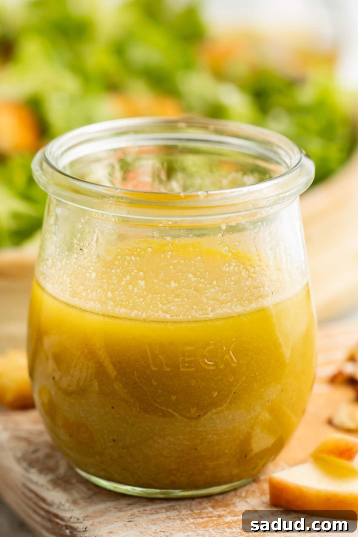 A second close-up view of the glass jar of vivid green apple cider vinaigrette on a wooden cutting board, surrounded by fresh green apple slices, with a blurred backdrop of a healthy salad.