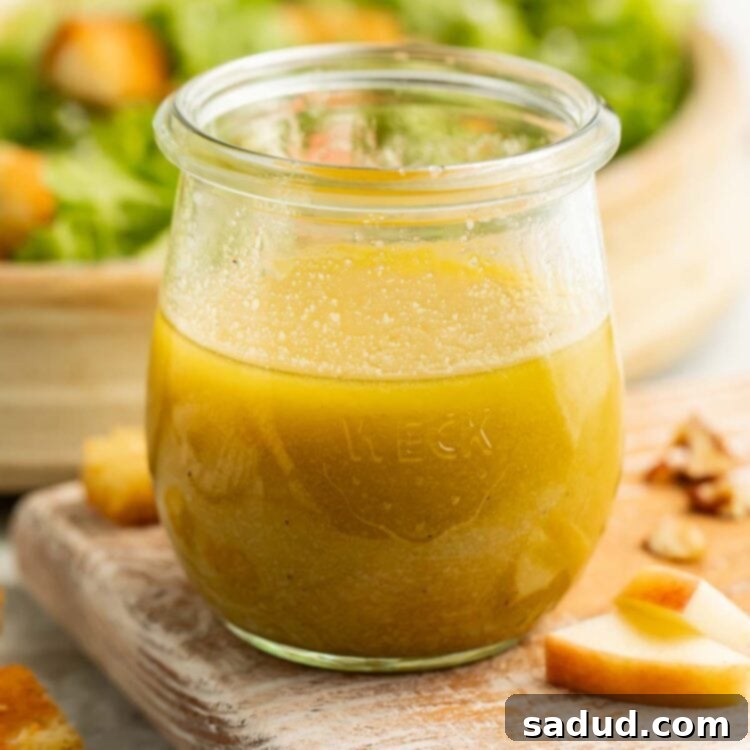 A glass jar of green apple cider vinaigrette on a cutting board surrounded by pieces of apple with a green salad in the background.